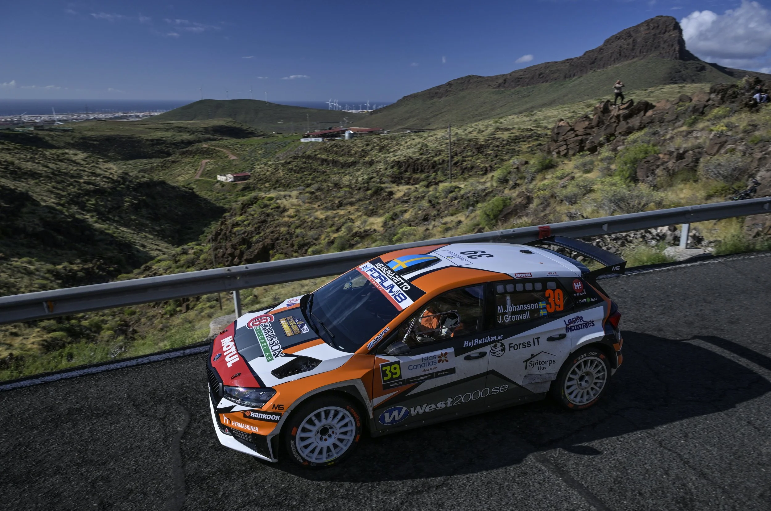 A rally car racing on a winding mountain road with scenic green hills and mountains in the background under a clear blue sky.