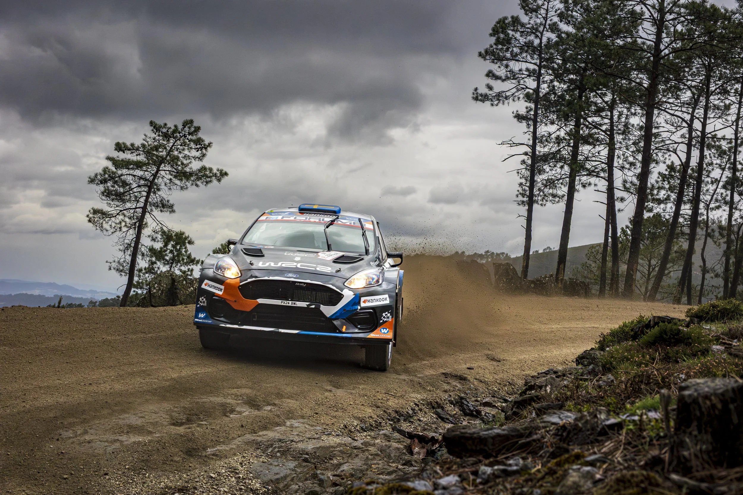 A rally car racing on a dirt road through a forested area with dark stormy clouds overhead.