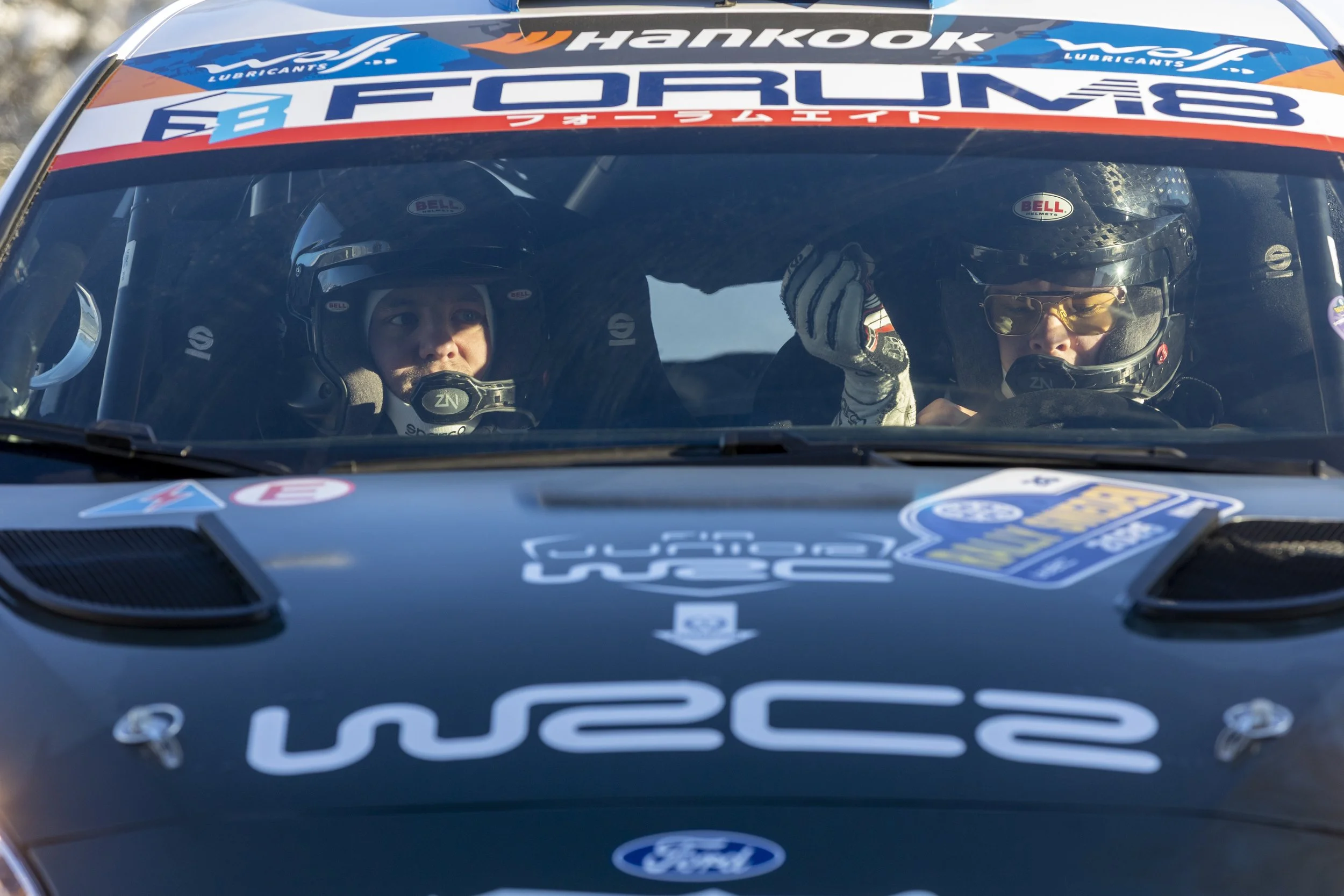 A racing car with two drivers inside, both wearing helmets and racing suits, sitting in the car before a race or practice session, with various sponsorship stickers and logos on the car's windshield and hood.