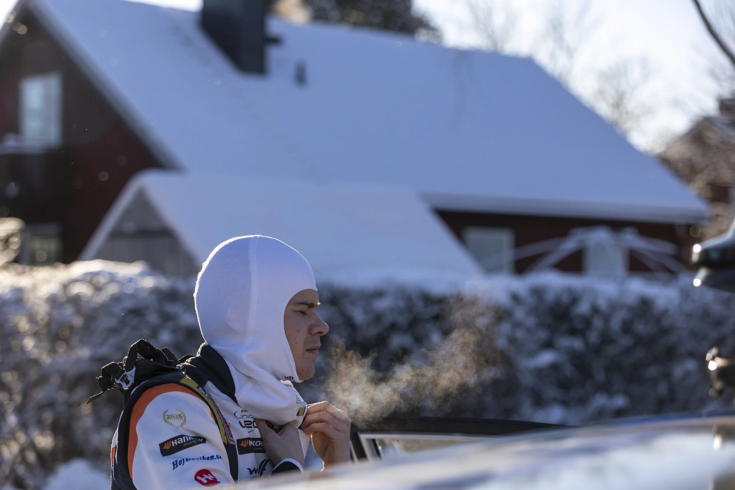 A woman dressed in winter sports gear, including a white hood and a jacket with sponsor logos, stands outdoors in a snowy landscape with a house and trees in the background.