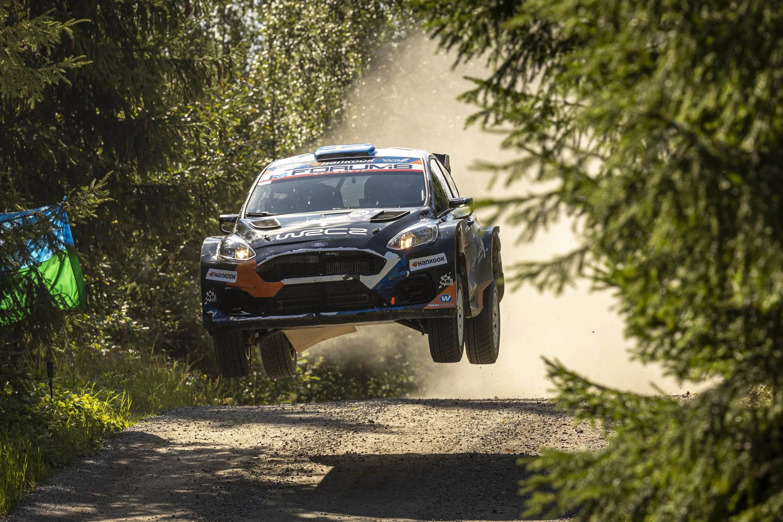 A rally car airborne over a dirt road in a wooded forest.