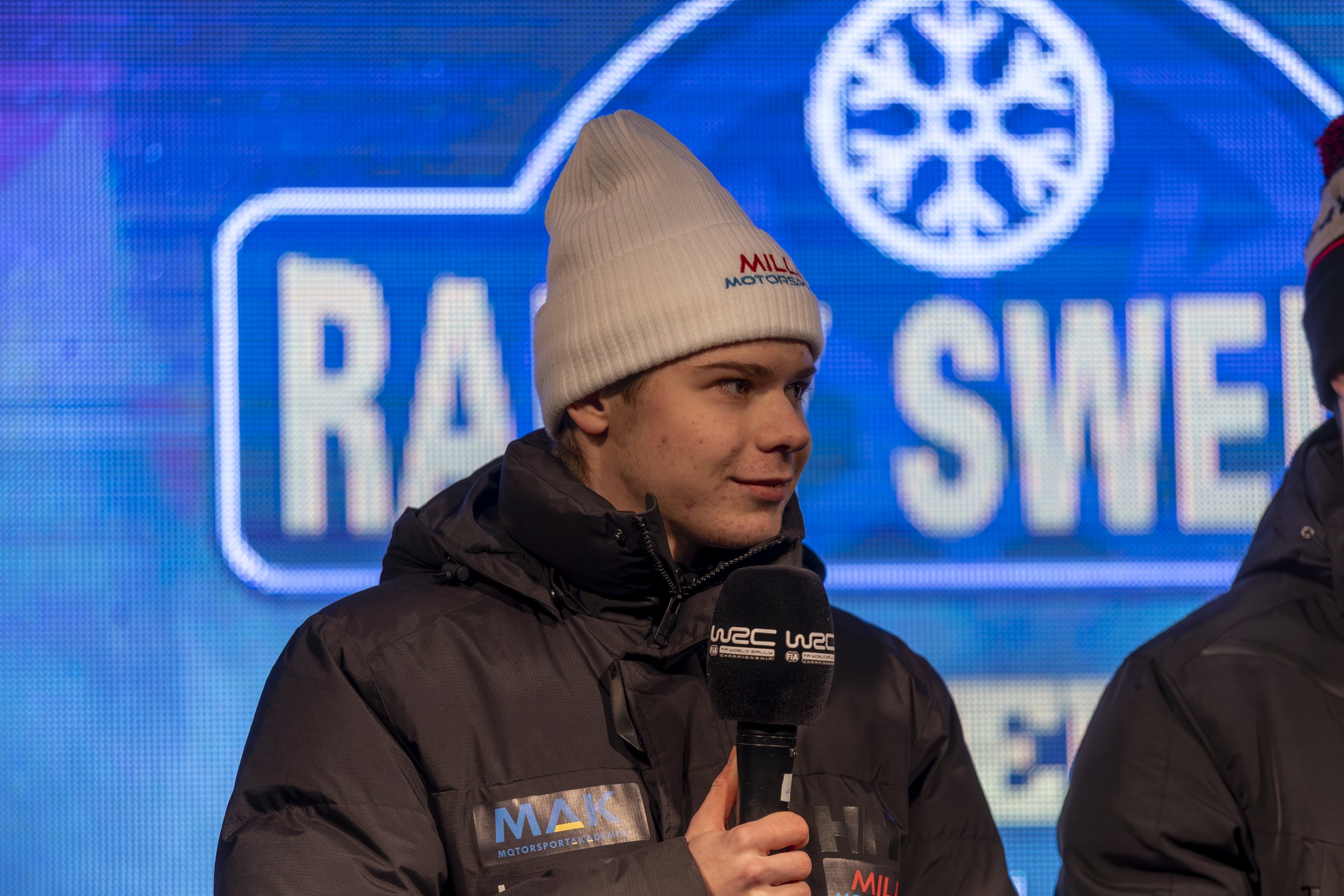 Young man in a white beanie and black jacket holding a microphone labeled 'WRC' during a racing event interview with a blue digital backdrop displaying the 'FIA World Rally Championship' logo.
