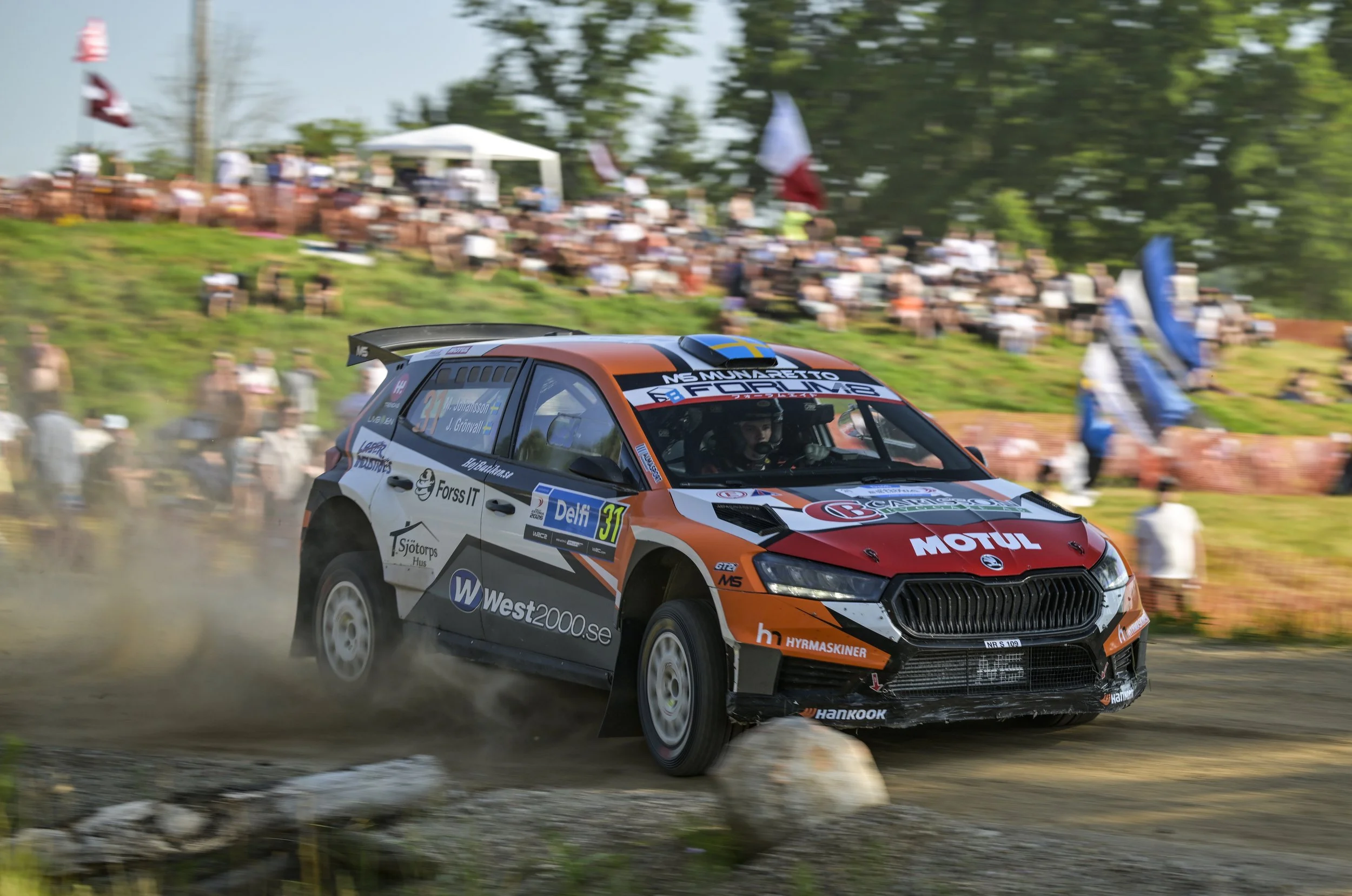 A rally car in motion on a dirt race track with a crowd of spectators watching in the background.