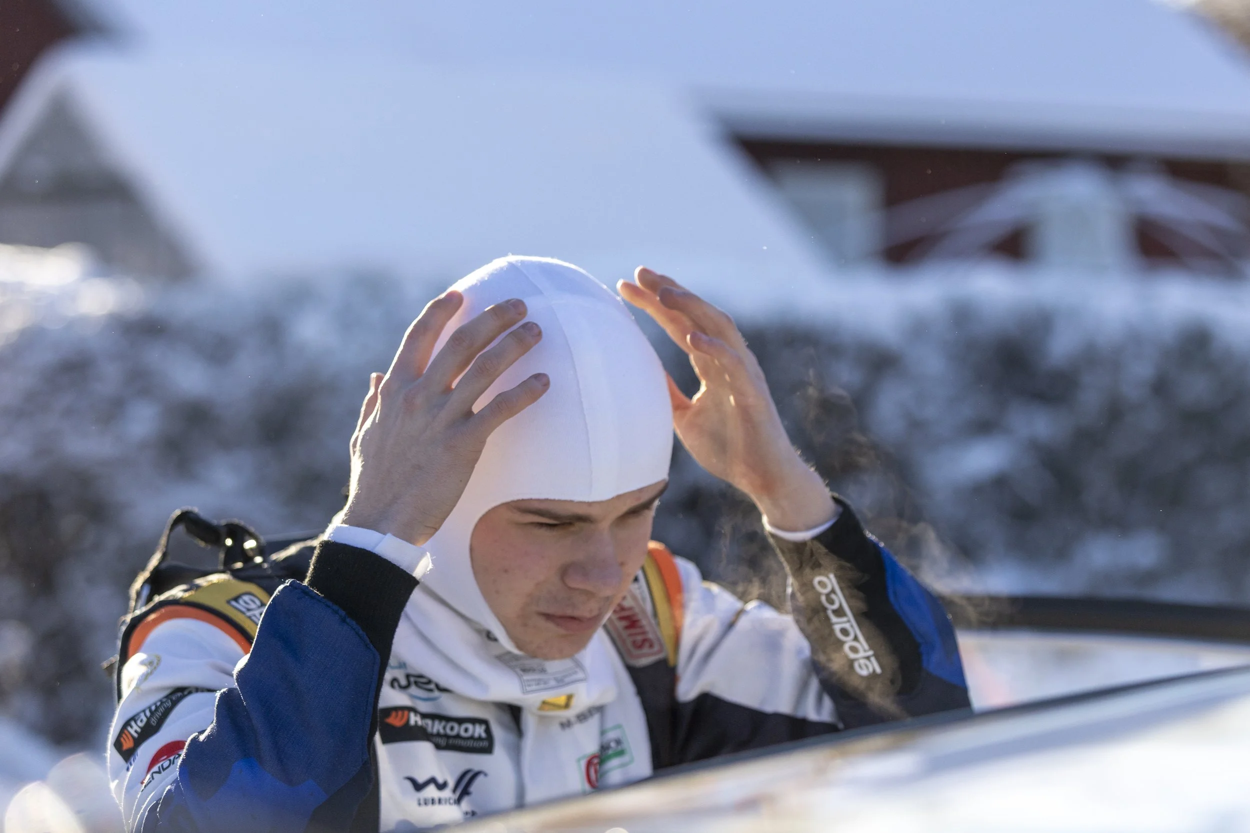 A young athlete in a racing suit adjusting his white racing helmet at a snowy outdoor location, with a blurred backdrop of snow and sky.