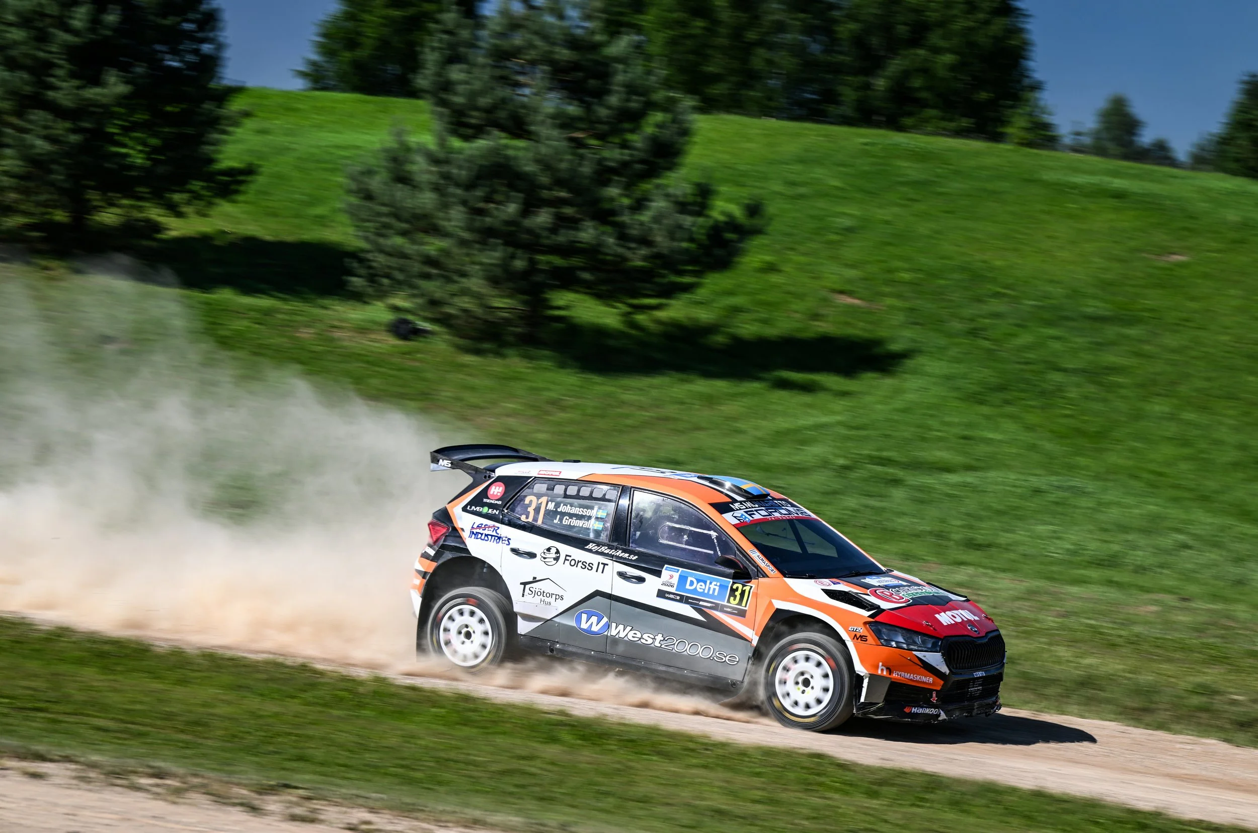 A rally car with number 31 speeds on a dirt road, kicking up dust, with a grassy hillside and trees in the background.