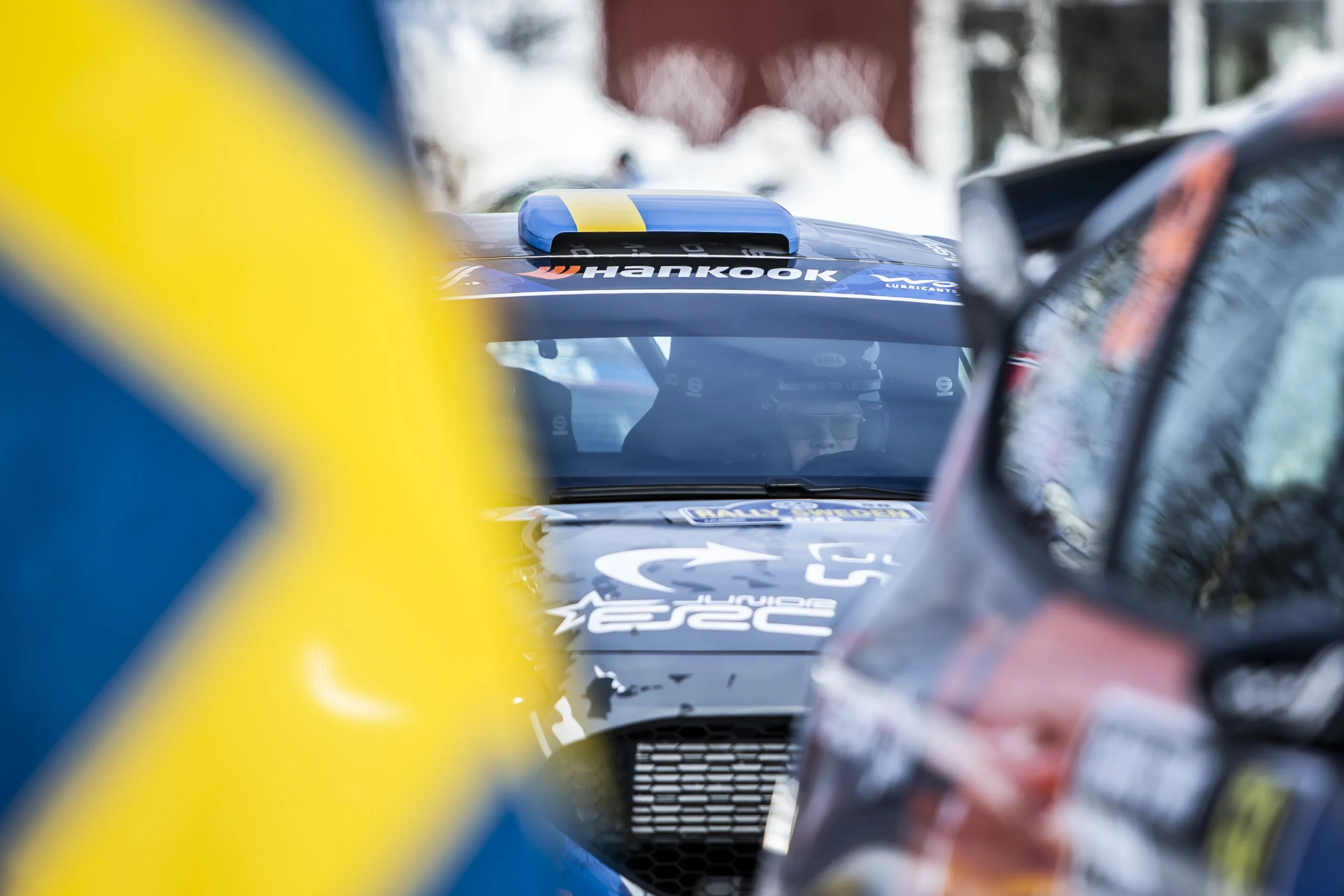 View through a car's windshield showing racing cars, including a blue car with Hankook branding, amidst a crowded scene with blurred figures and snow in the background.