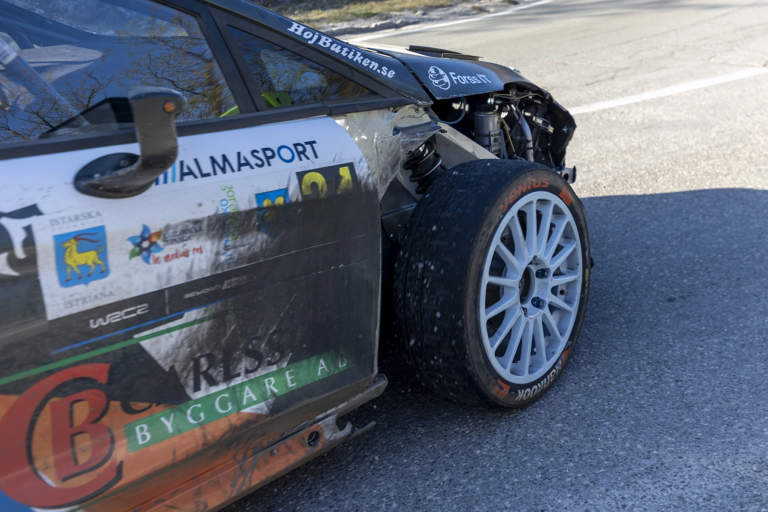 Close-up of a race car's side showing sponsor decals, a white wheel with a Goodyear tire, and part of the suspension, parked on a paved road.