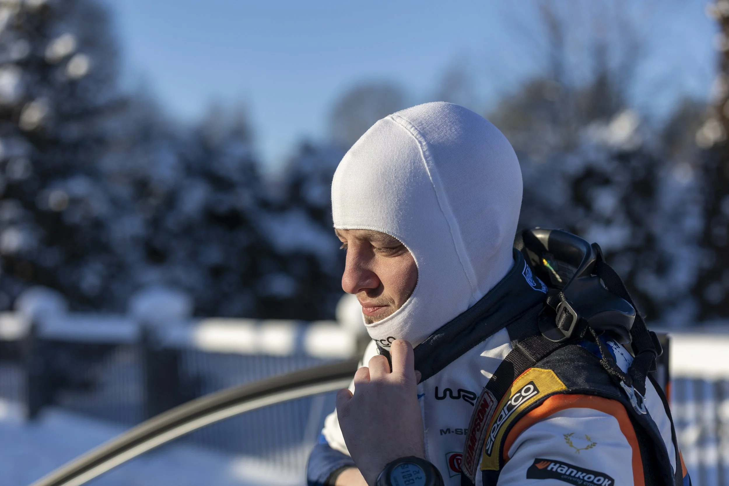 Close-up of a person in racing gear wearing a white hat outdoors in a snowy environment, preparing for a race.