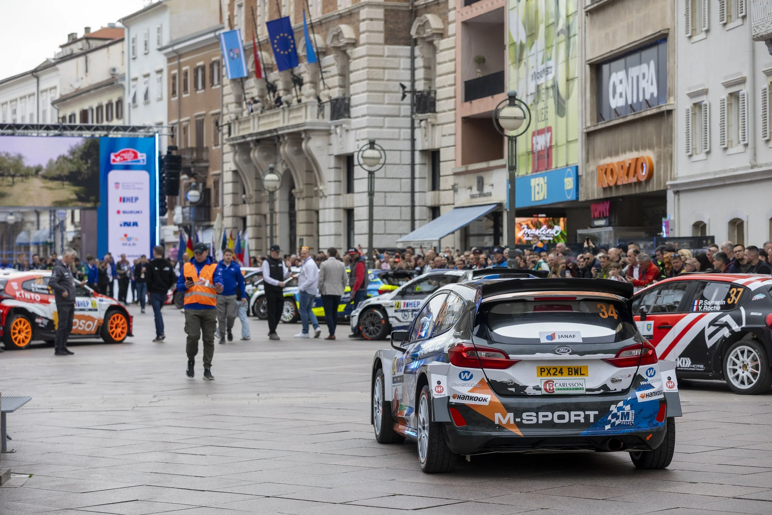 Race cars lining up in a city square with a crowd of spectators and event staff around them, historic buildings in the background, flags, and large screens.