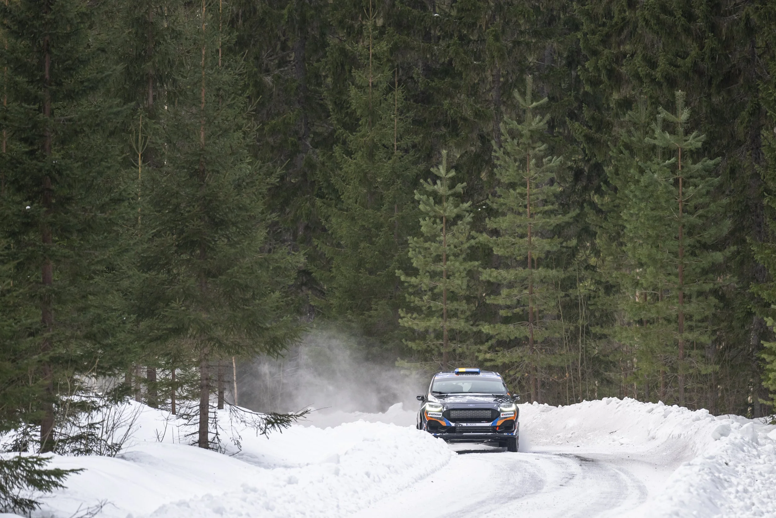 A car driving on a snow-covered forest road surrounded by tall pine trees, with snow piled along the roadside and smoke or mist in the background.