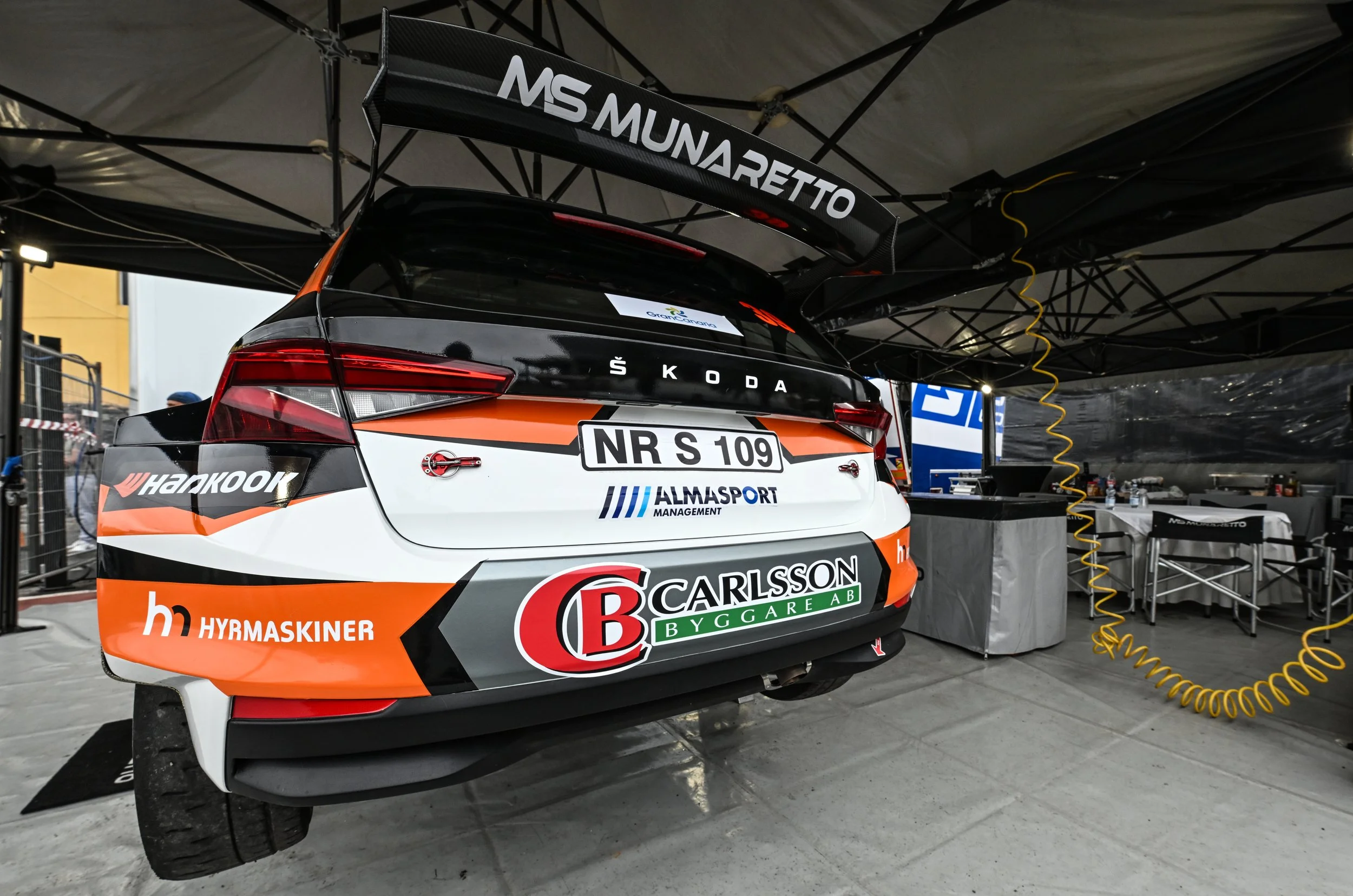 A race car in a service area under a canopy, with sponsor and team logos, and a large rear wing with 'MS Mundarett' branding.