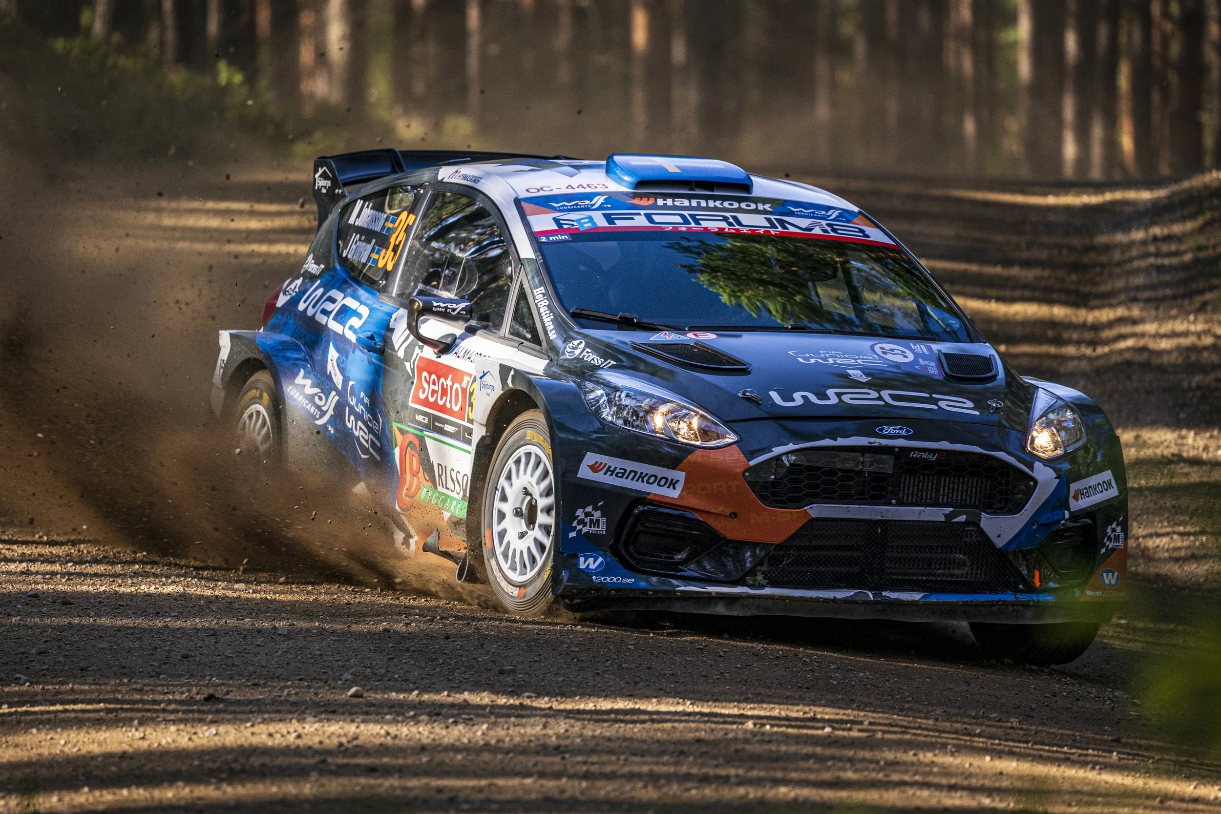 A blue and black rally car racing on a dirt forest road kicking up dust.
