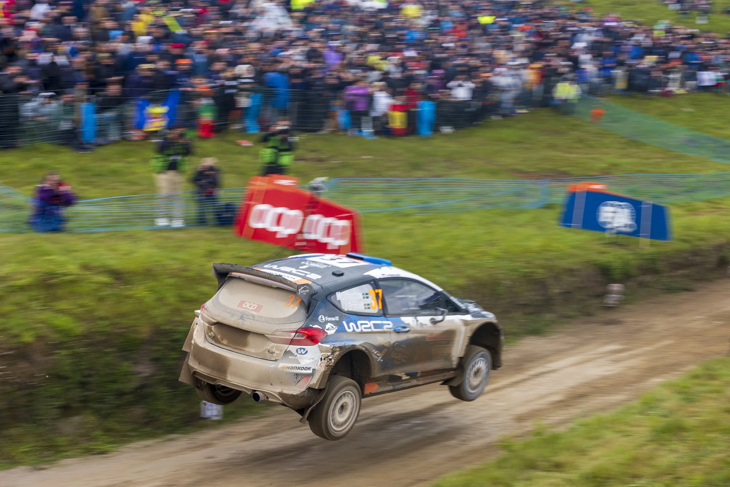 A dirt rally car with the number 37 jumps off a dirt track, kicking up dust, during a rally race. In the background, a crowd of spectators watches the event, separated by fencing, with some dressed in colorful clothing and a few security personnel pr