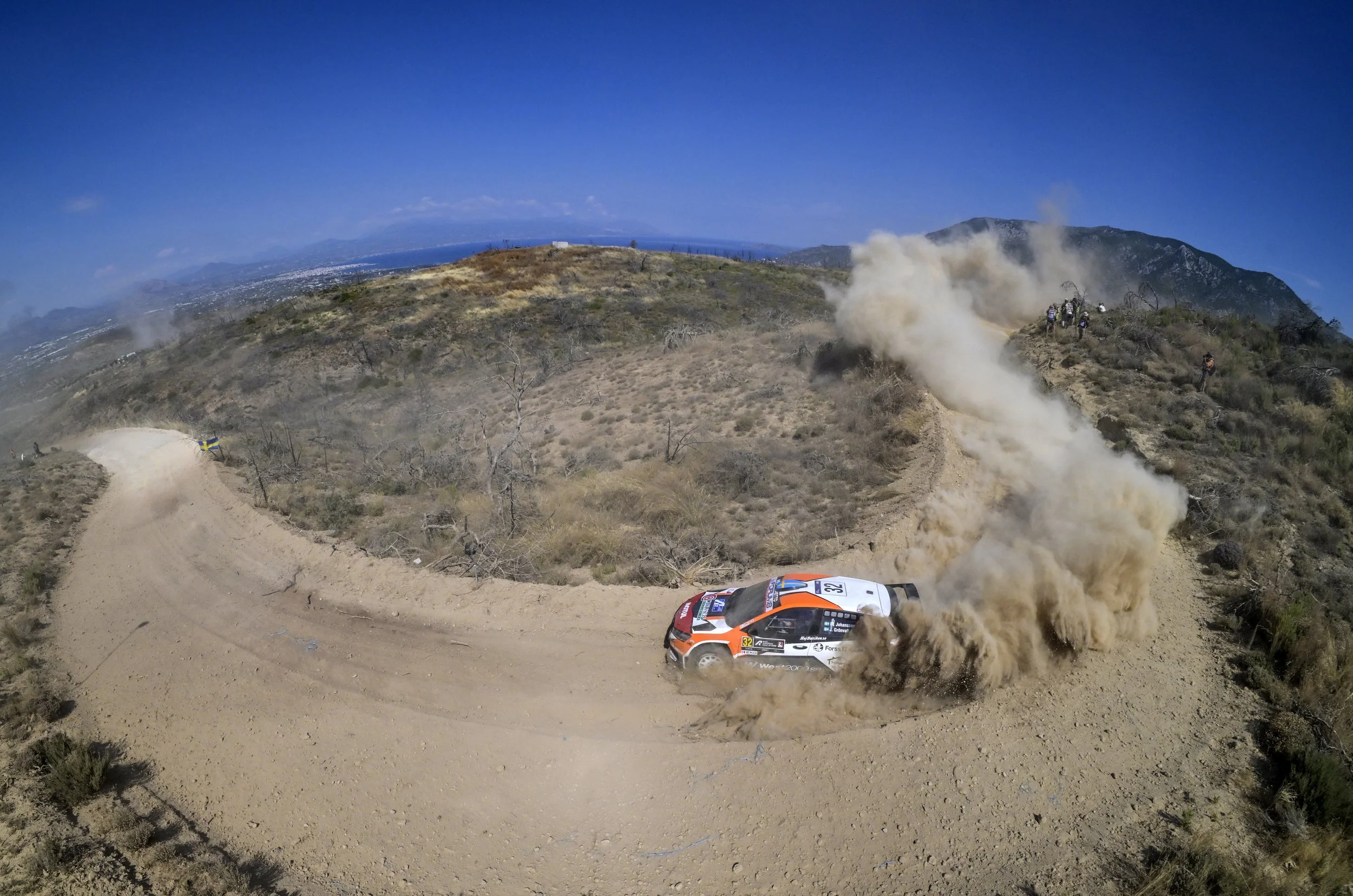 A rally car racing on a dirt trail in a mountainous area, kicking up dust, with a small group of spectators and a flag nearby, under a clear blue sky.