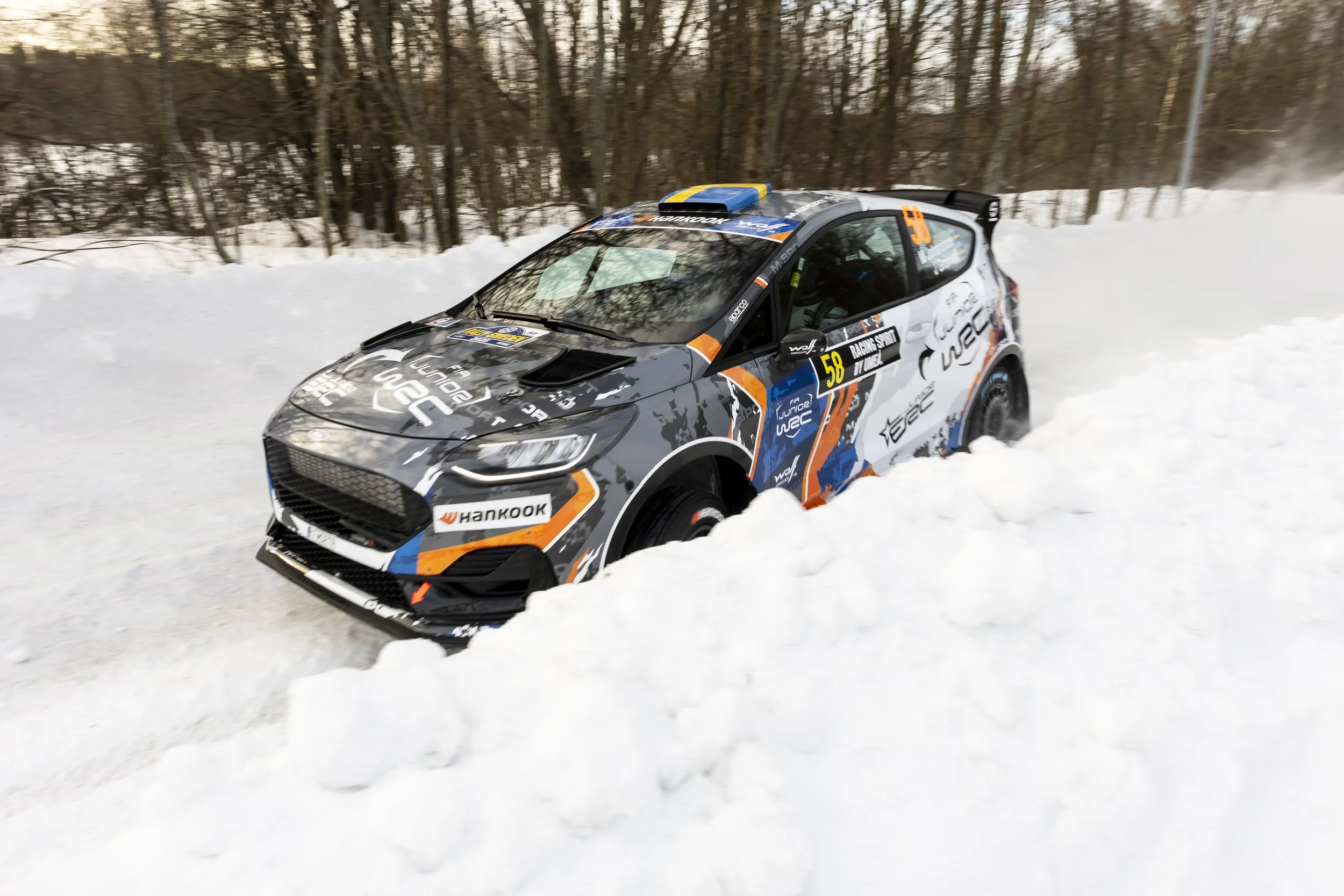 A rally car speeding through snow on a winter forested road, kicking up snow dust behind it.