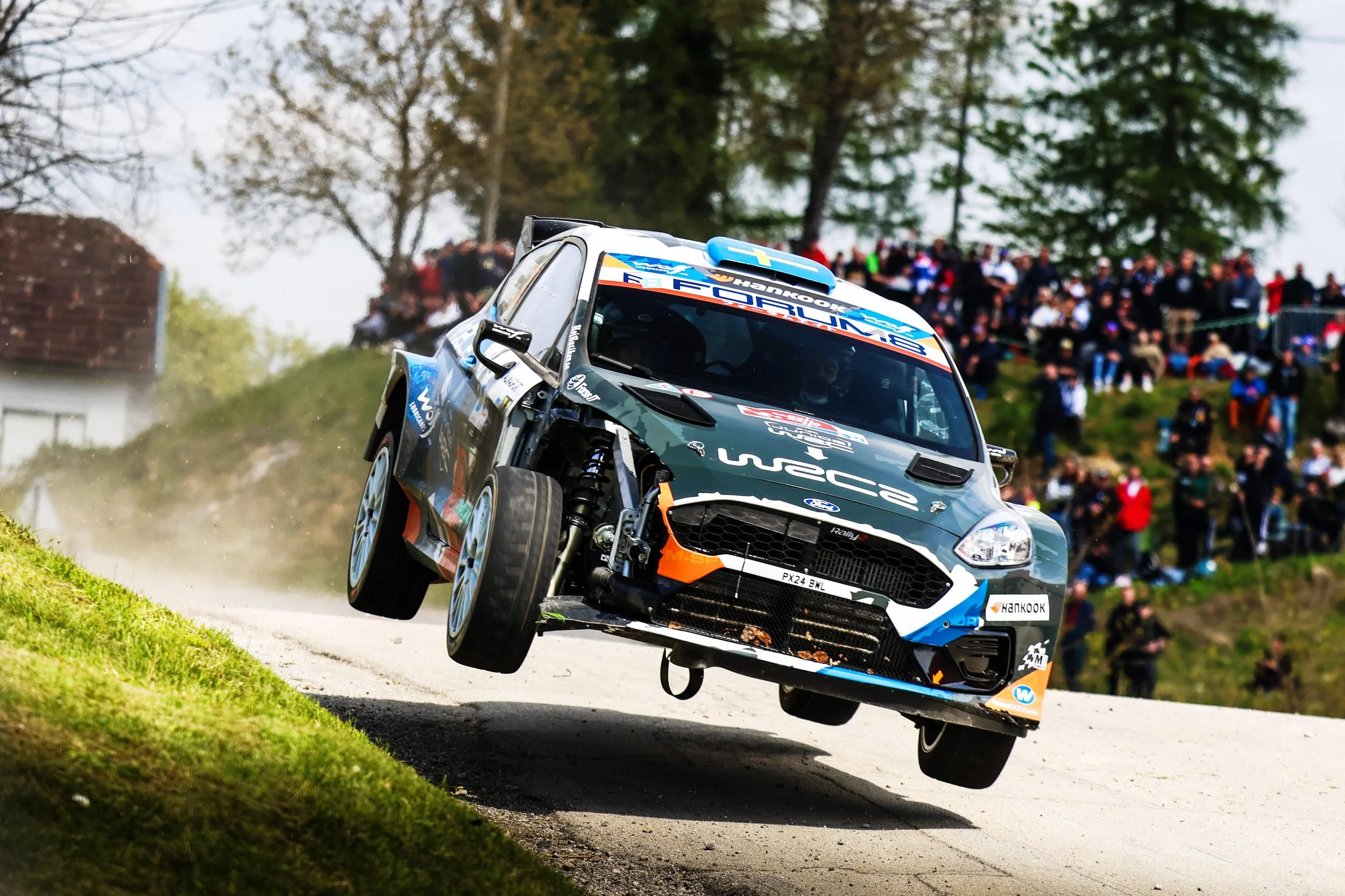 A rally car jumping off a dirt road during a race, with a crowd of spectators watching from the background.