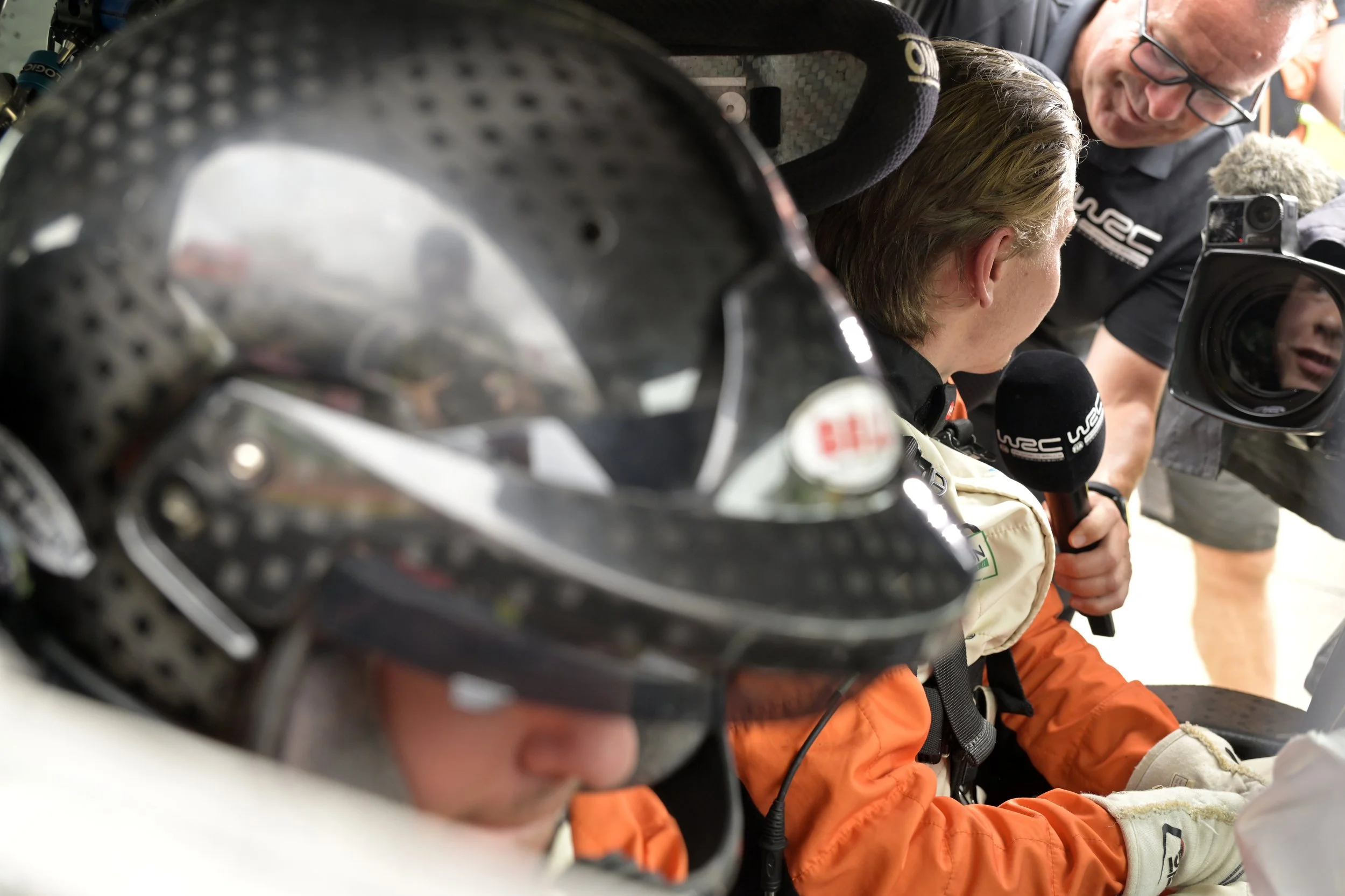 Two men and a woman wearing racing gear, with one holding a microphone, are gathered around a racing car, likely discussing the vehicle.