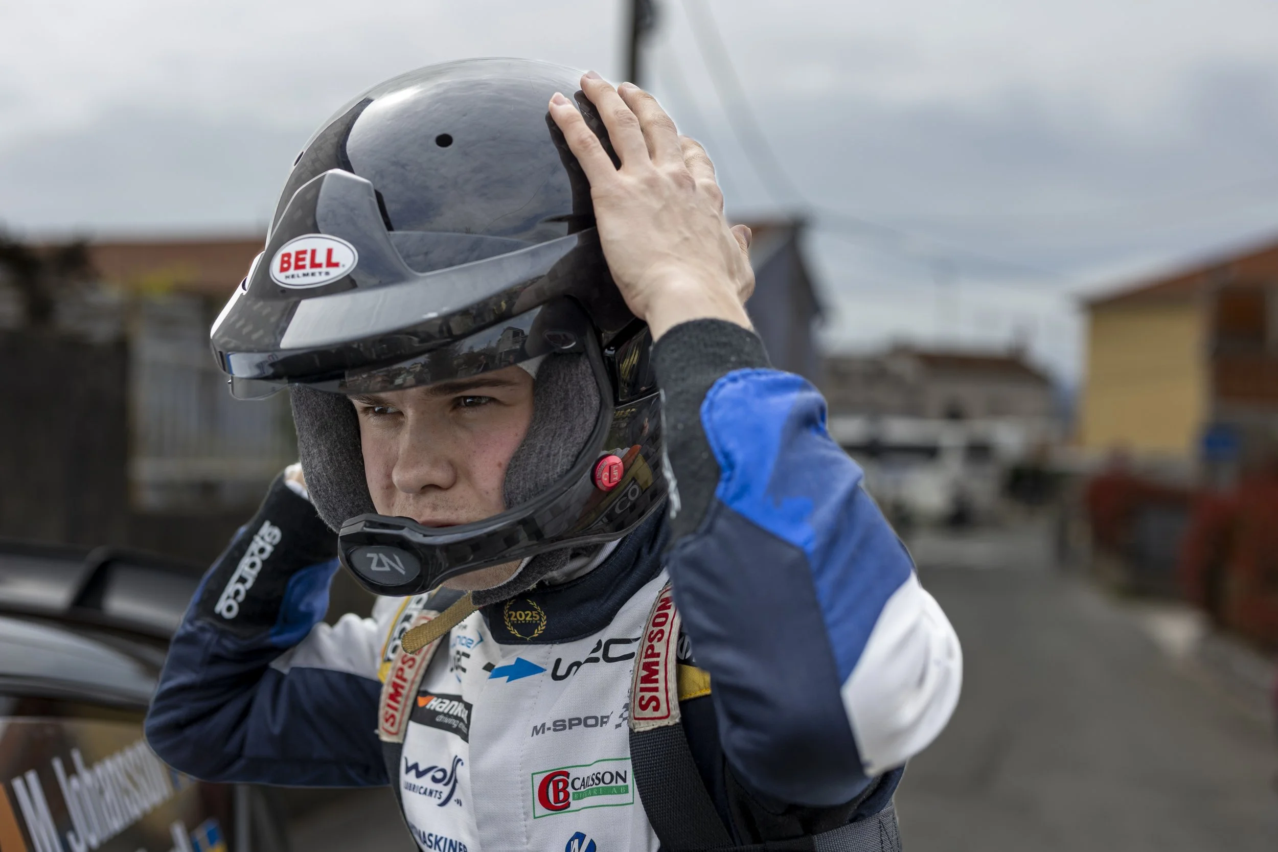 A race car driver adjusting his helmet before a race, wearing a blue and white racing suit with various sponsor logos, outdoors with a cloudy sky in the background.