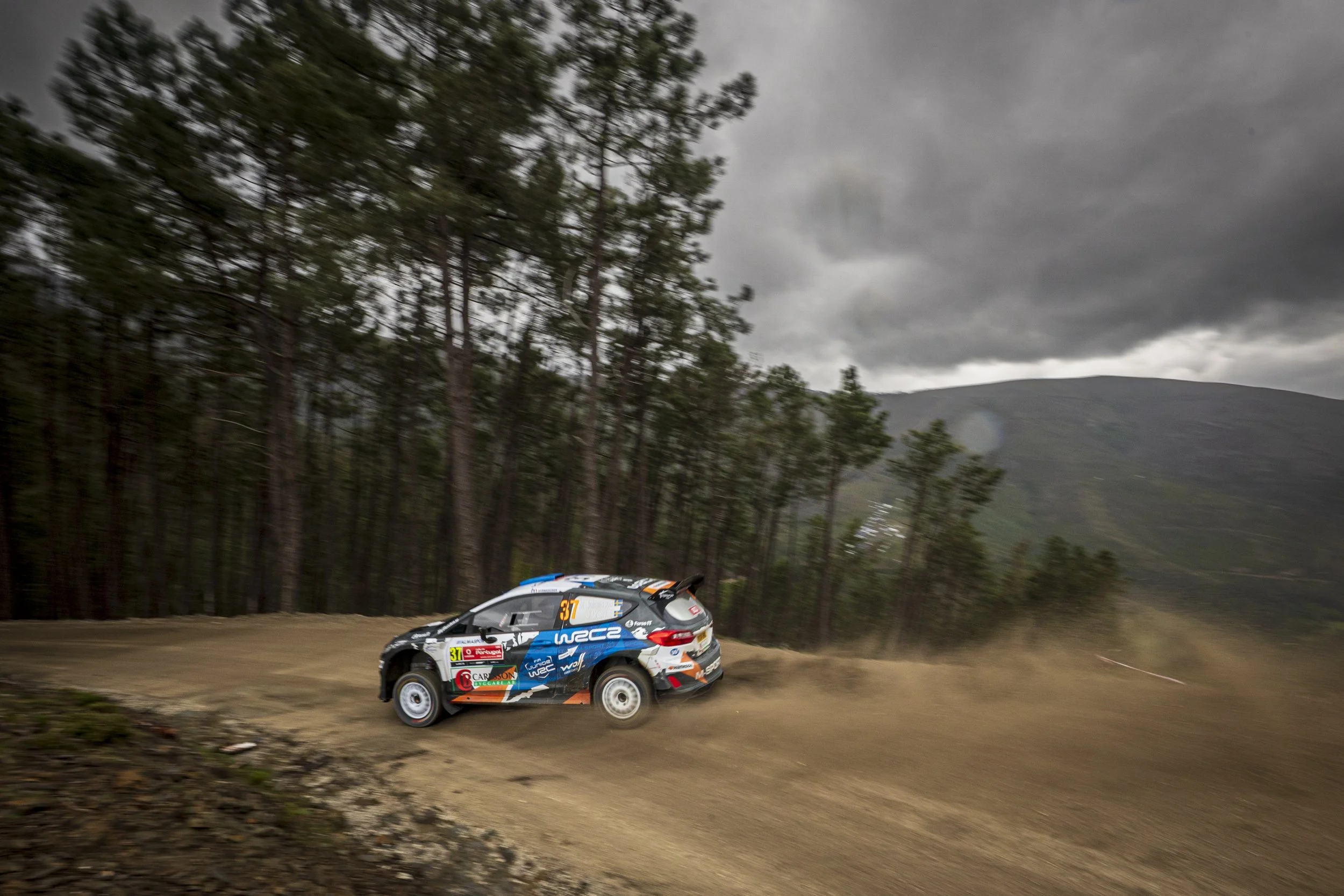 A rally car racing on a dirt road with trees on the side, mountains in the background, and dark cloudy skies overhead.