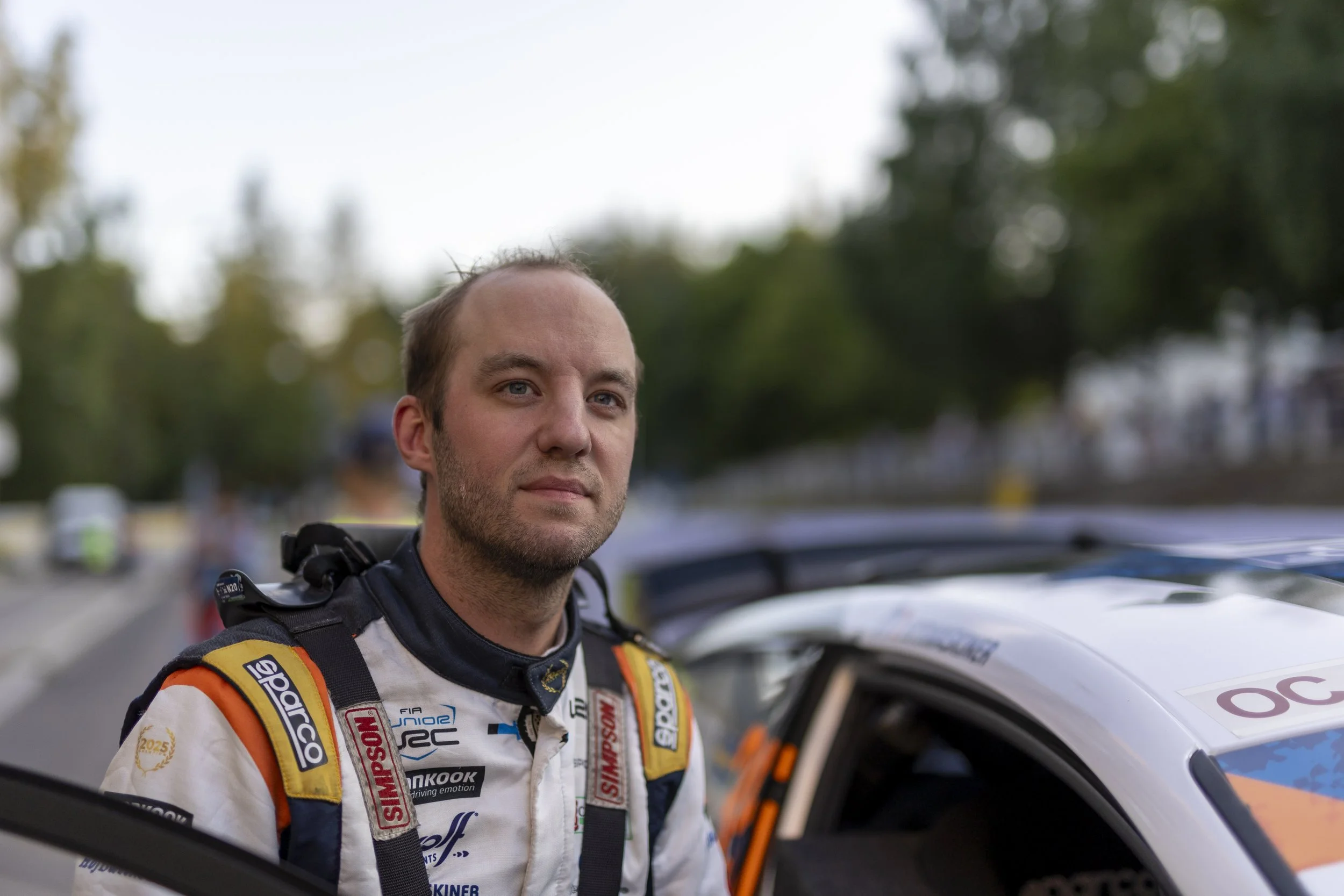 Close-up of a male race car driver in a racing suit standing outdoors next to his race car, with trees and sky in the background.
