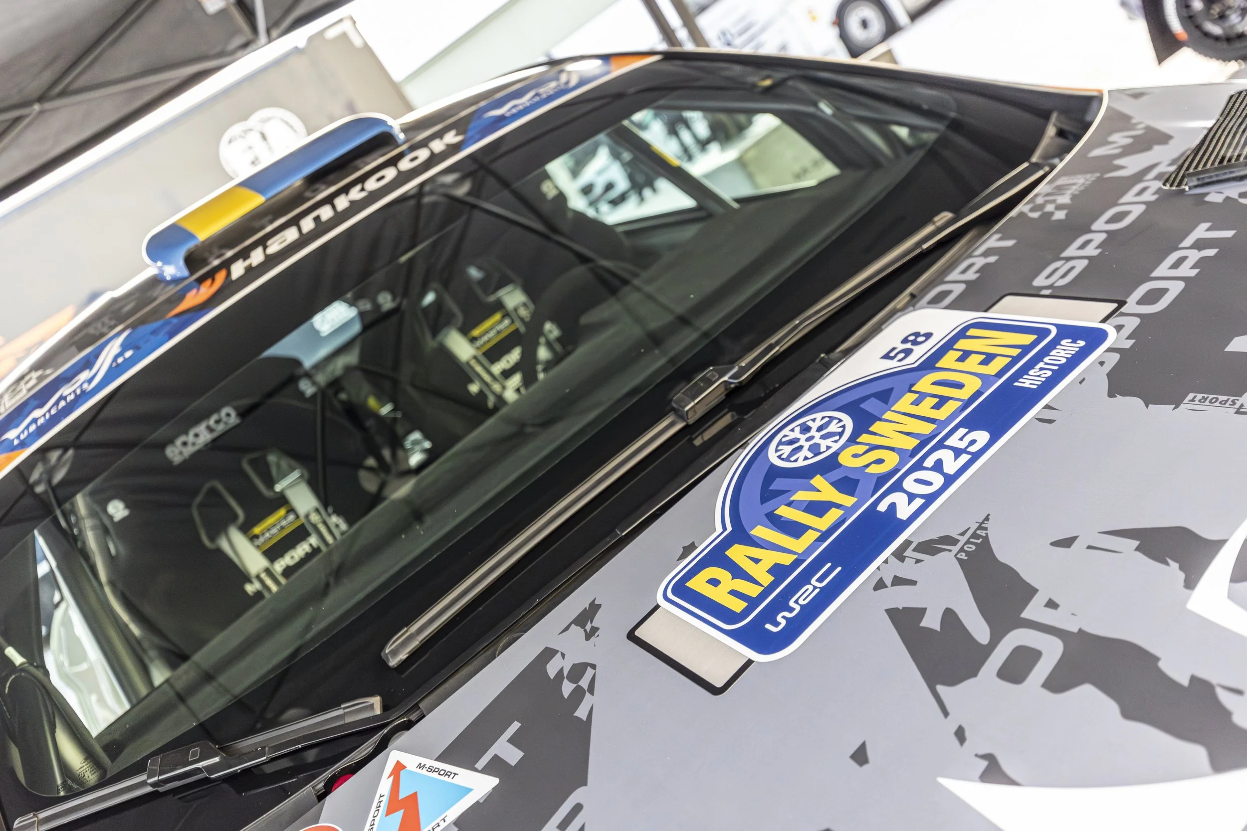 Close-up of a race car with sponsor stickers, including a large racing sticker on the hood, set in a garage or workshop.