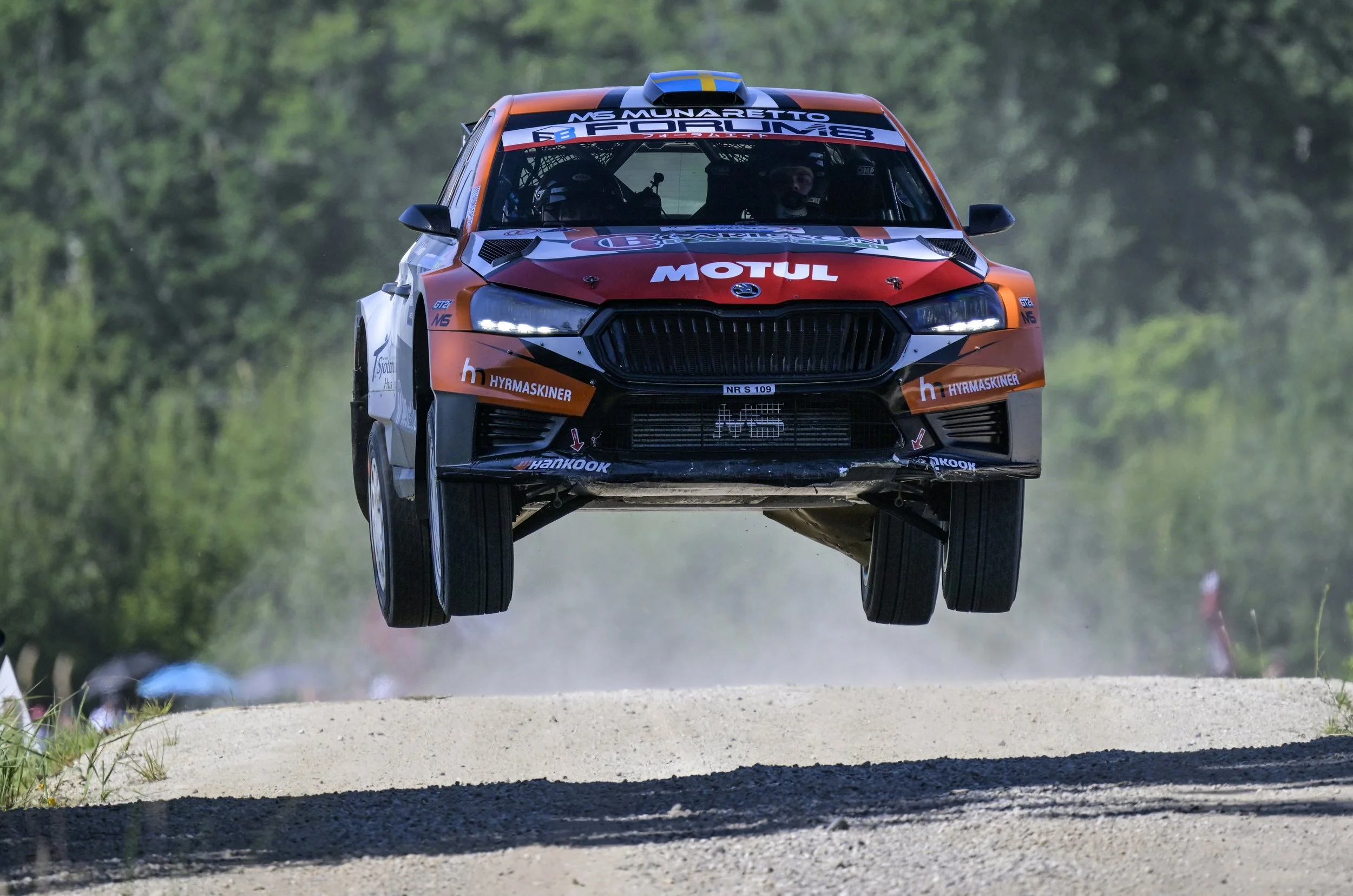 A rally car flying in the air during a race on a dirt track, with a green forest background.
