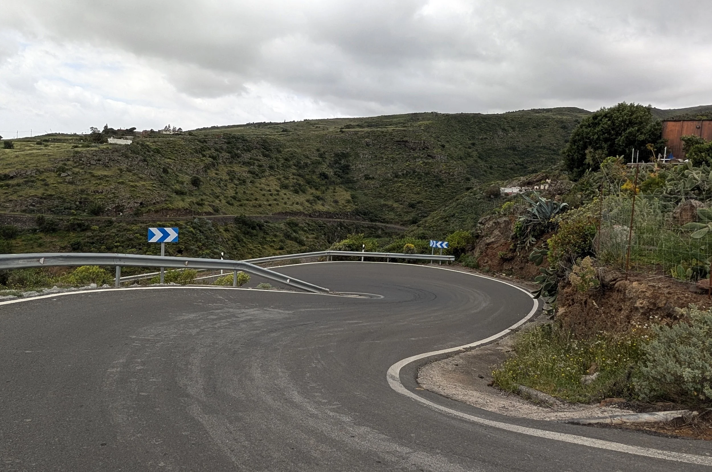 A winding mountain road with a metal guardrail and blue directional arrows, surrounded by green hillside and cloudy sky.