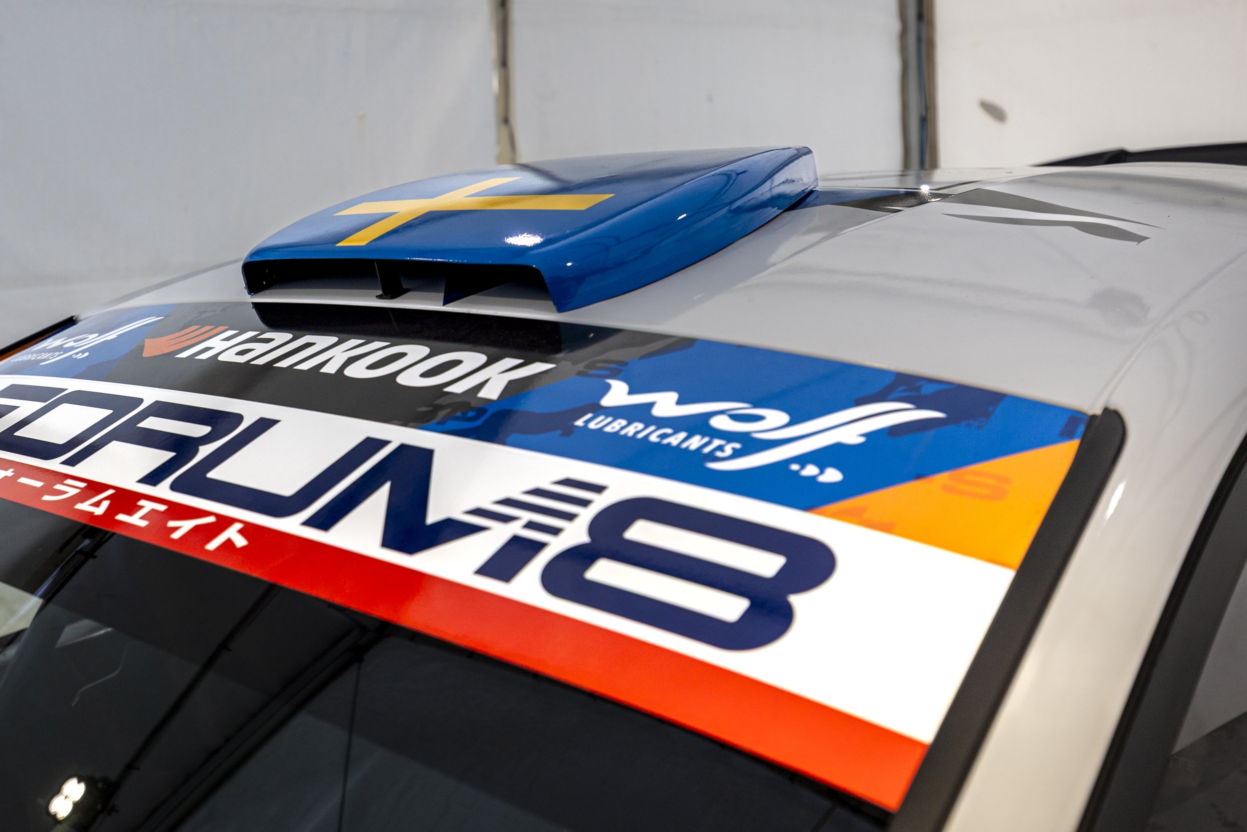 Close-up of racing car hood with stickers, including blue air intake with yellow cross, and various racing sponsor logos.