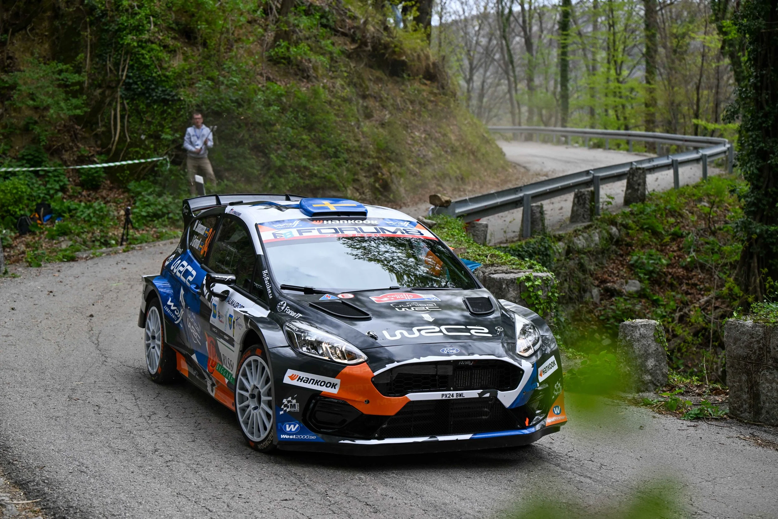 A rally car driving on a narrow, winding gravel road through a lush, green forest with trees and bushes on both sides.