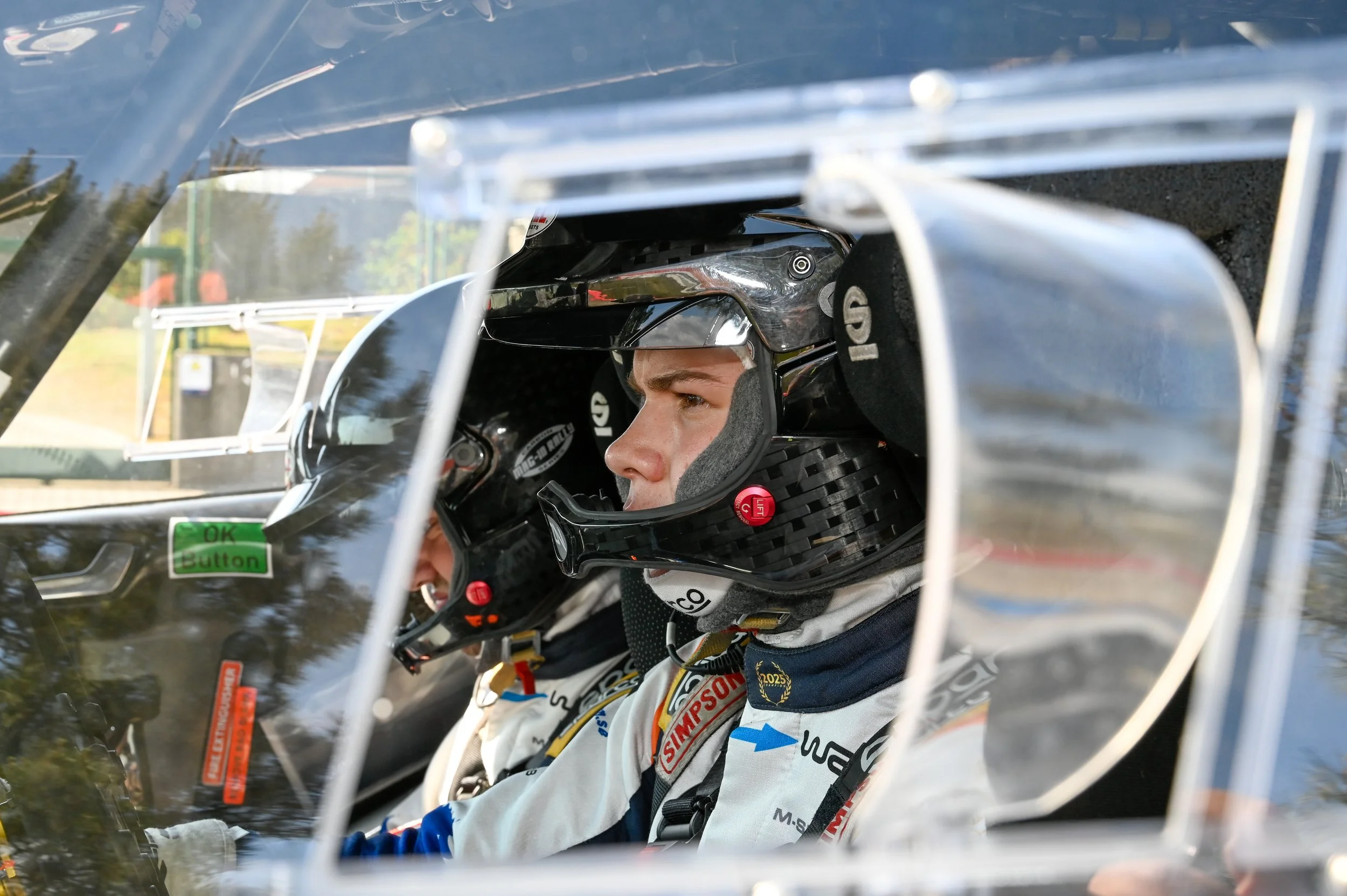 Race car driver wearing a helmet inside a race car cockpit, seen through the windshield.