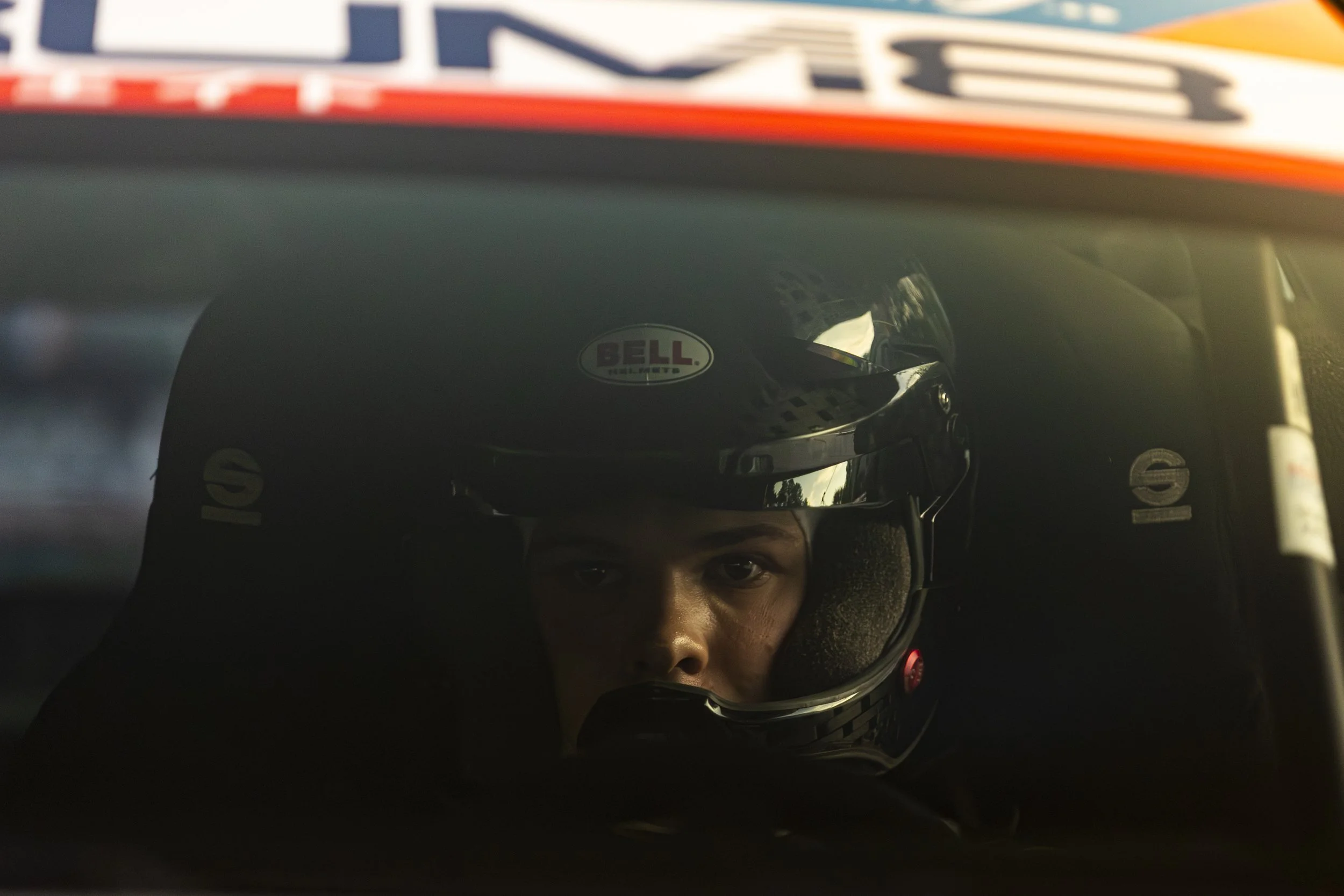 A person wearing a racing helmet peeking out from under the helmet's visor, with a red and white helmet sticker visible on the forehead area.
