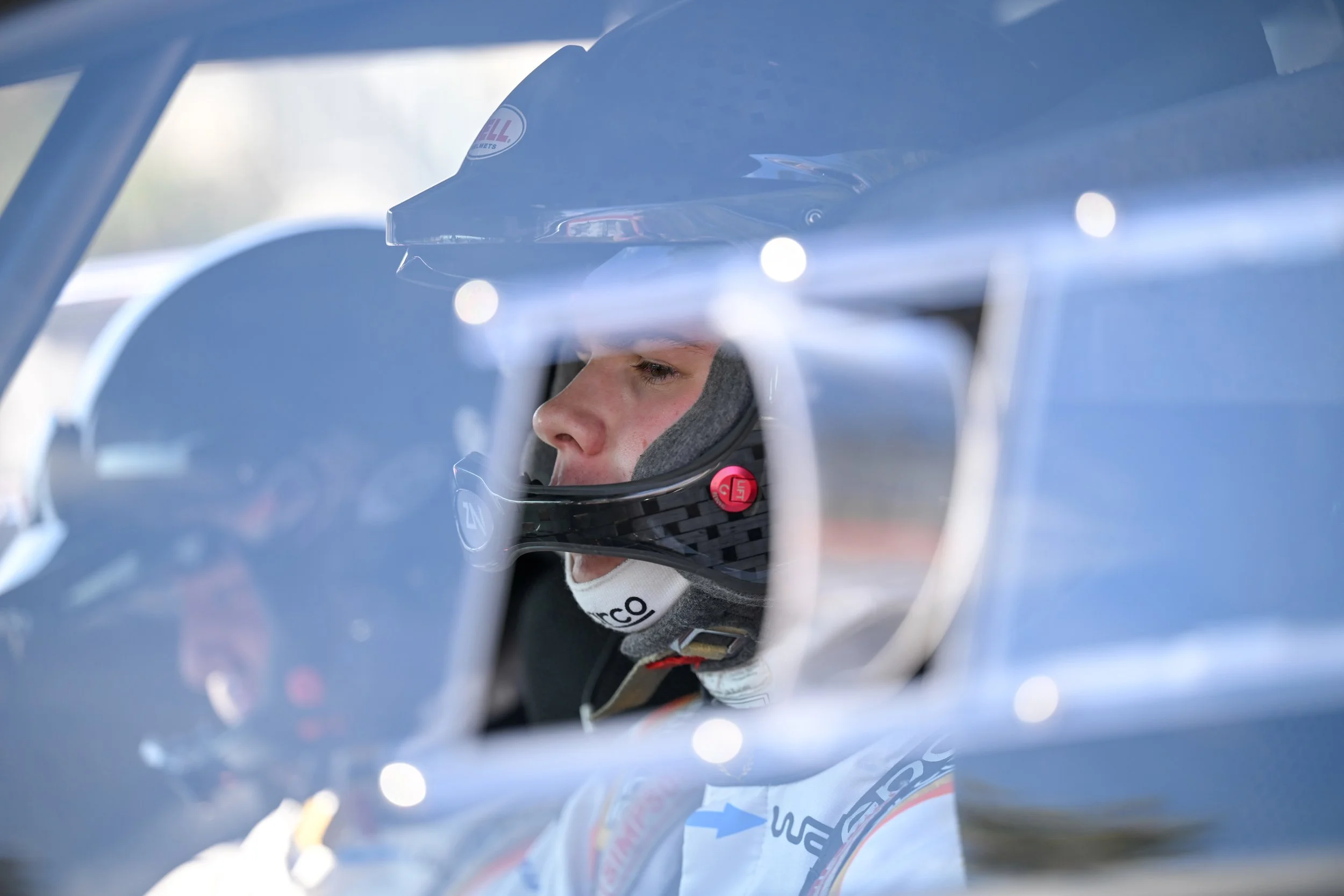 Close-up of a race car driver wearing a helmet, seen through the car's window, with the helmet facing sideways and the driver's face partially visible.