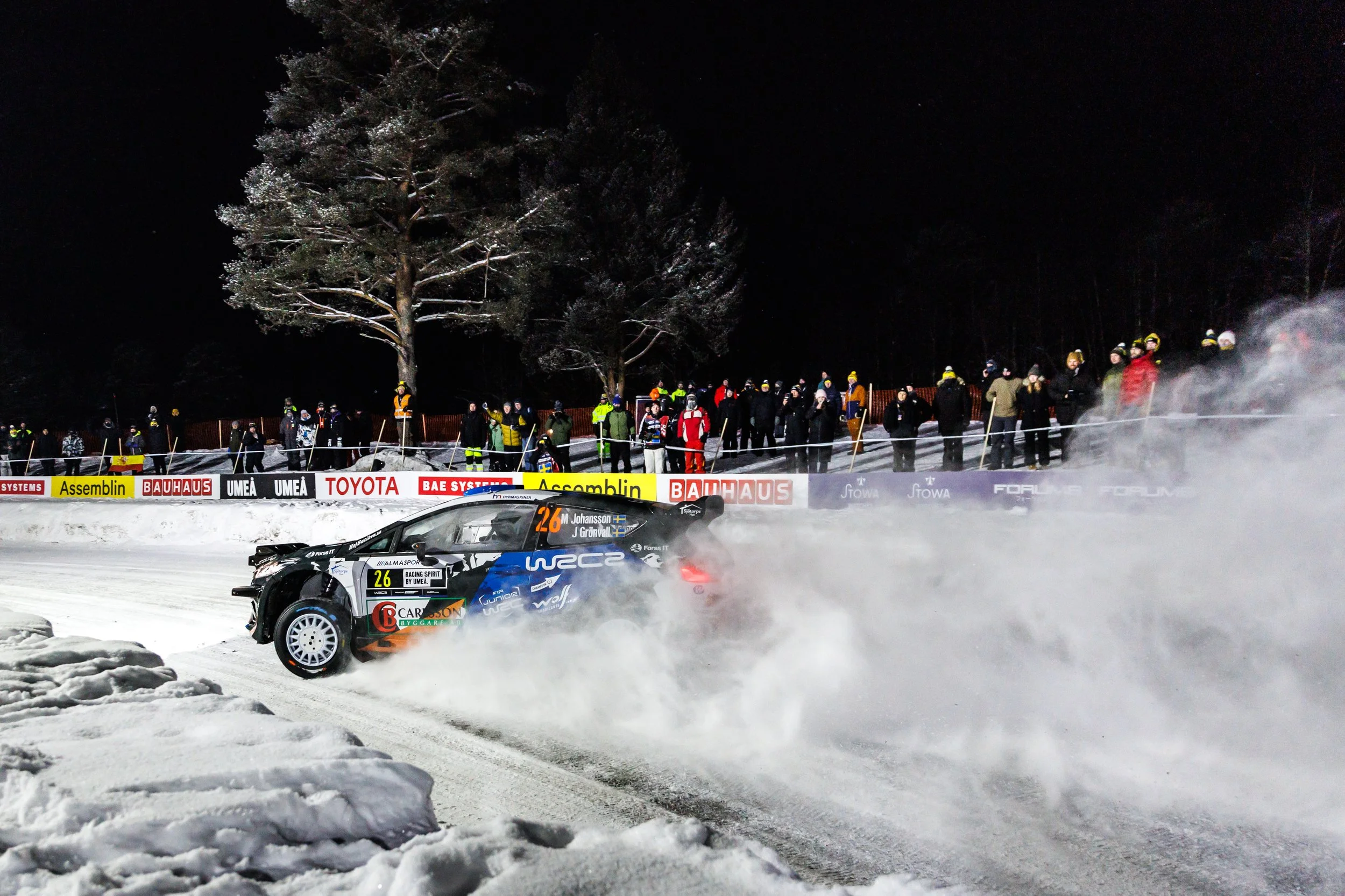 A black rally car with the number 26 on the side is racing on a snowy track at night, creating snow dust behind it. Spectators, some wearing winter clothing and helmets, stand along the track behind a safety barrier, watching the race.