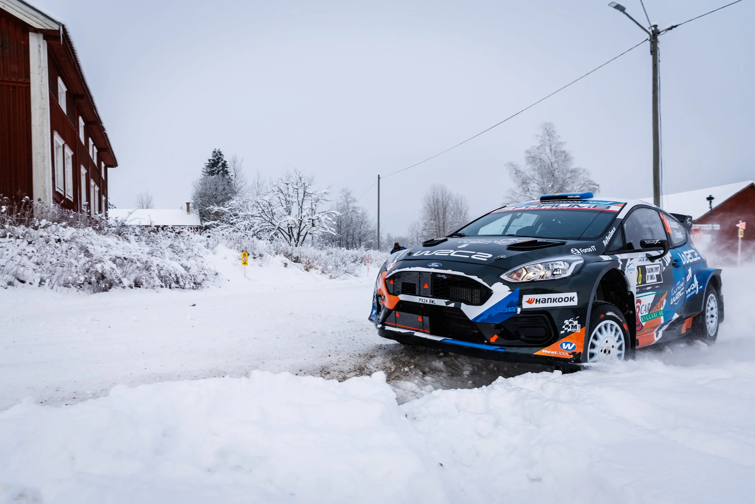 A black and blue rally car driving on a snow-covered road in a winter landscape with trees and houses, with snow on the ground and overhanging branches.