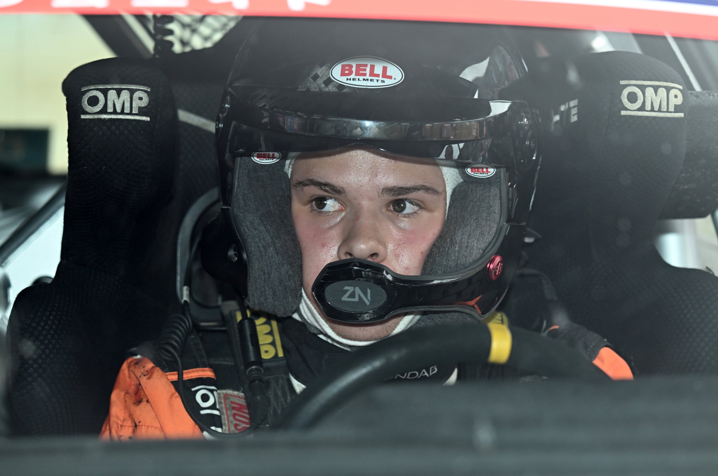 Race car driver wearing a Bell helmet and racing suit, sitting inside a race car, looking slightly upward.
