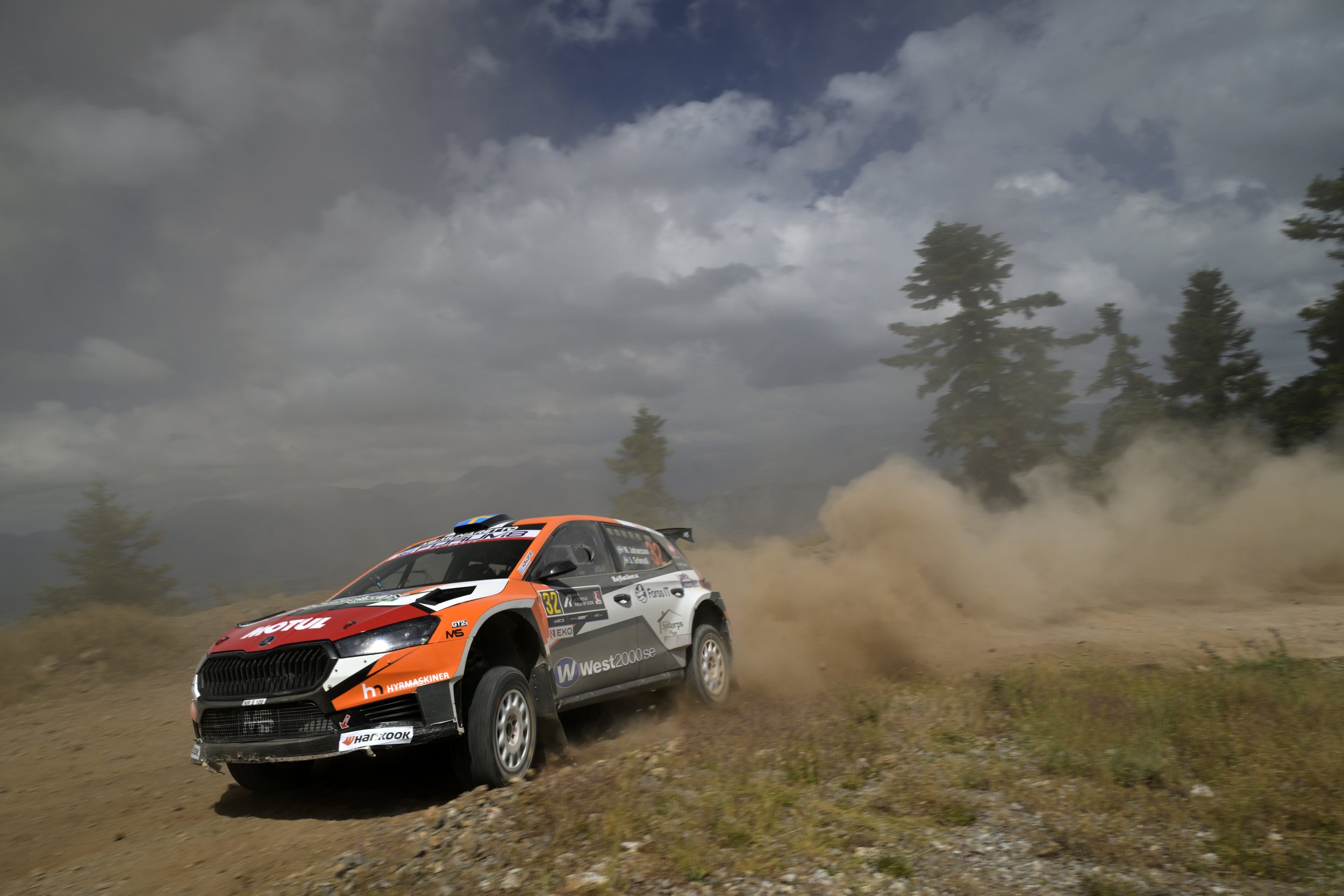 A rally car speeding along a dirt road with dust clouds and trees in the background, under a cloudy sky.