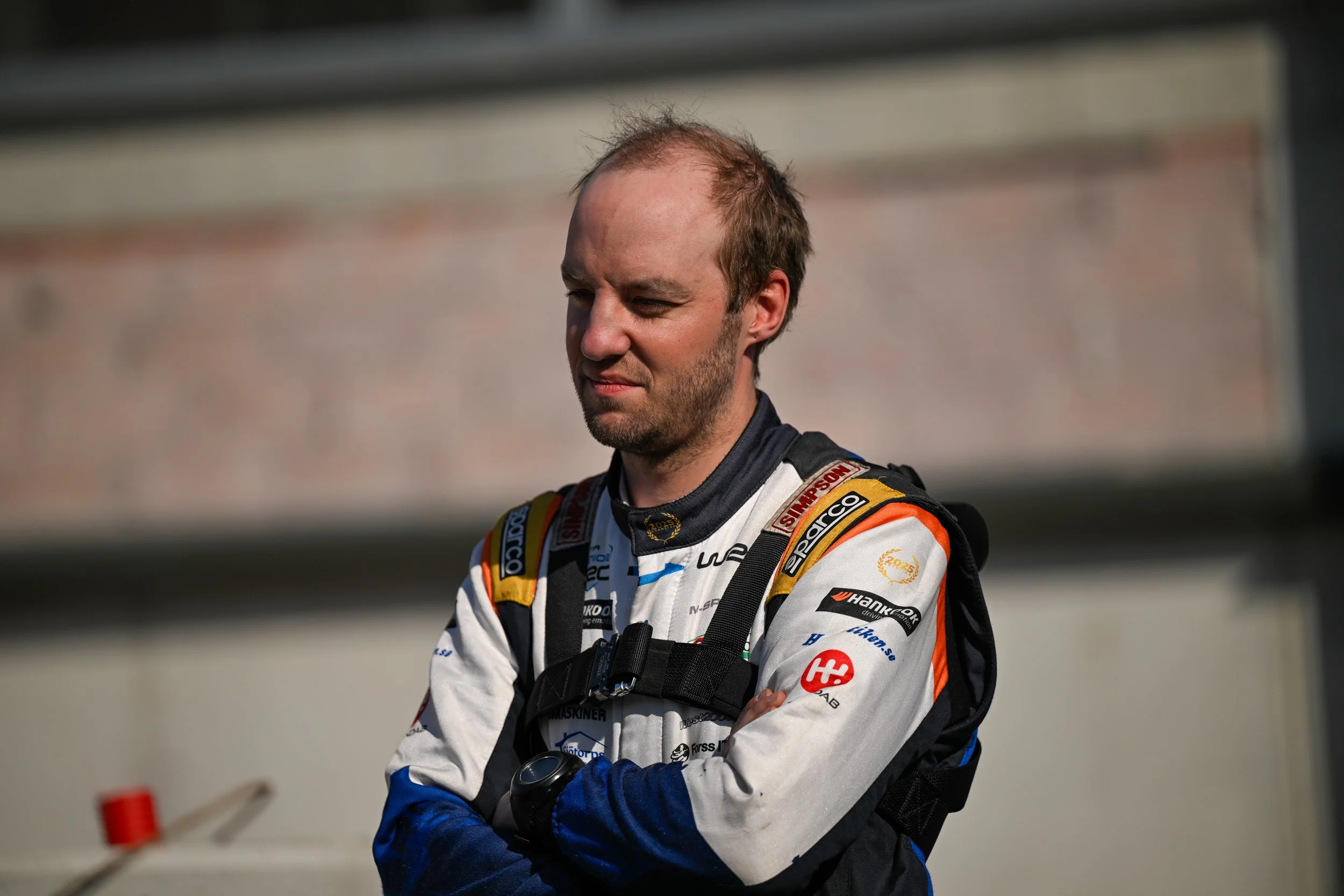A male race car driver wearing a racing suit and gloves, standing with his arms crossed, outdoors in sunlight.