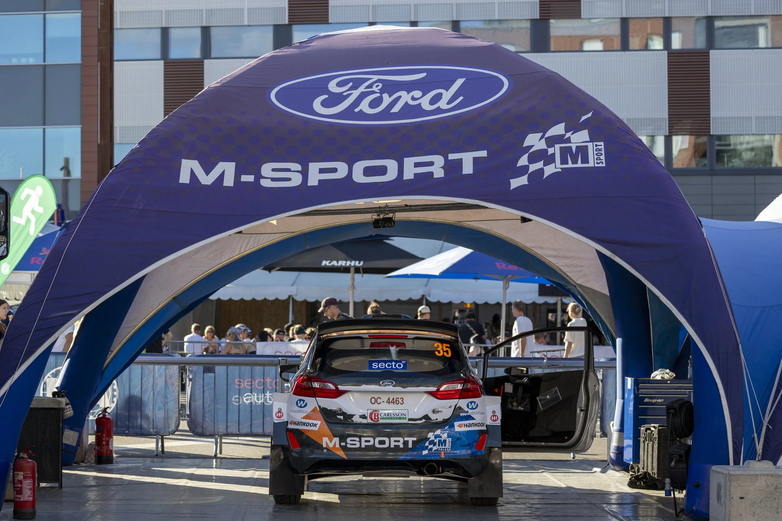 A racing car under a blue Ford M-Sport canopy at a motorsport event, with spectators and other event setups in the background.