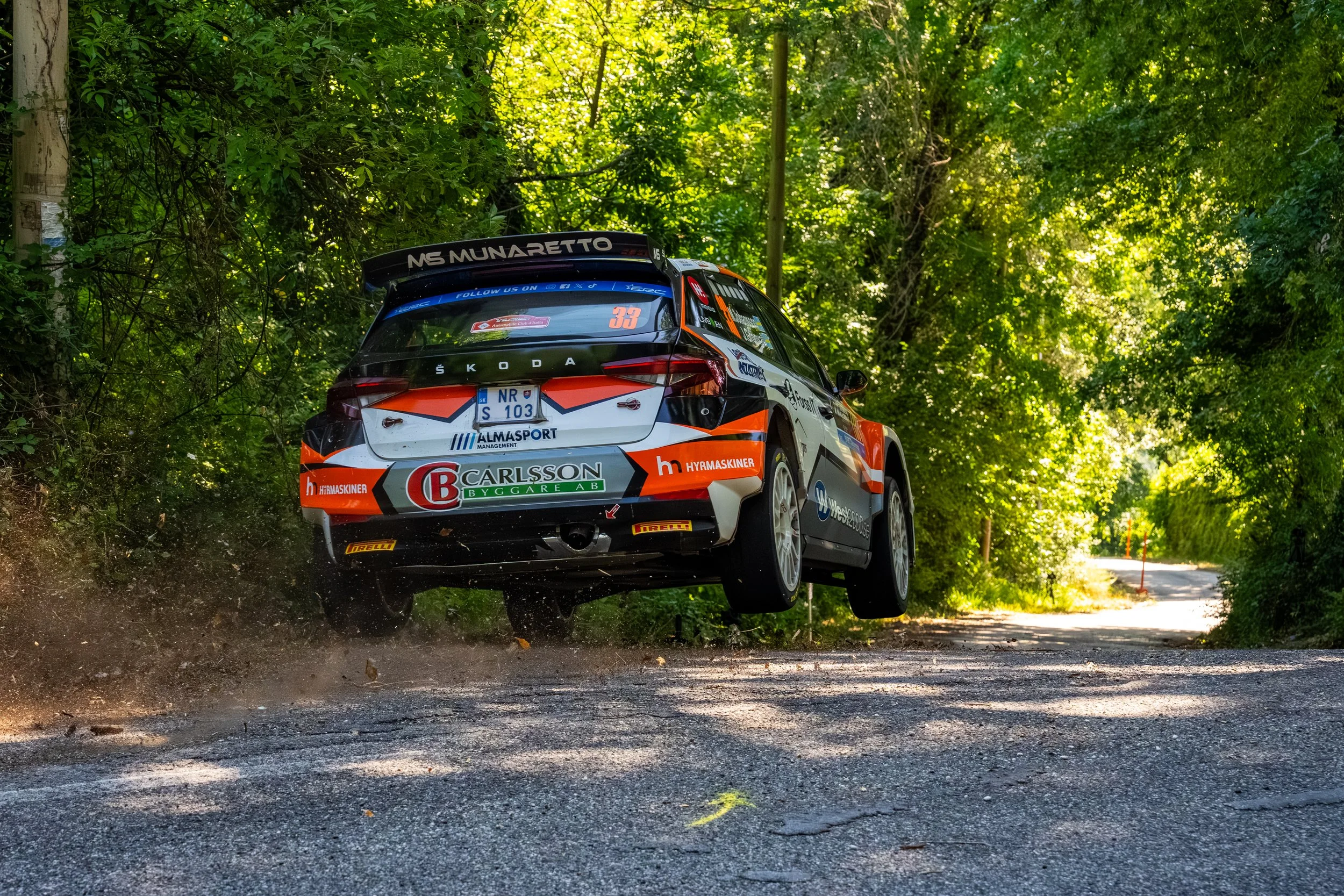 A rally car jumps over a dirt road in a forested area with lush green trees, dust and dirt flying beneath the vehicle.