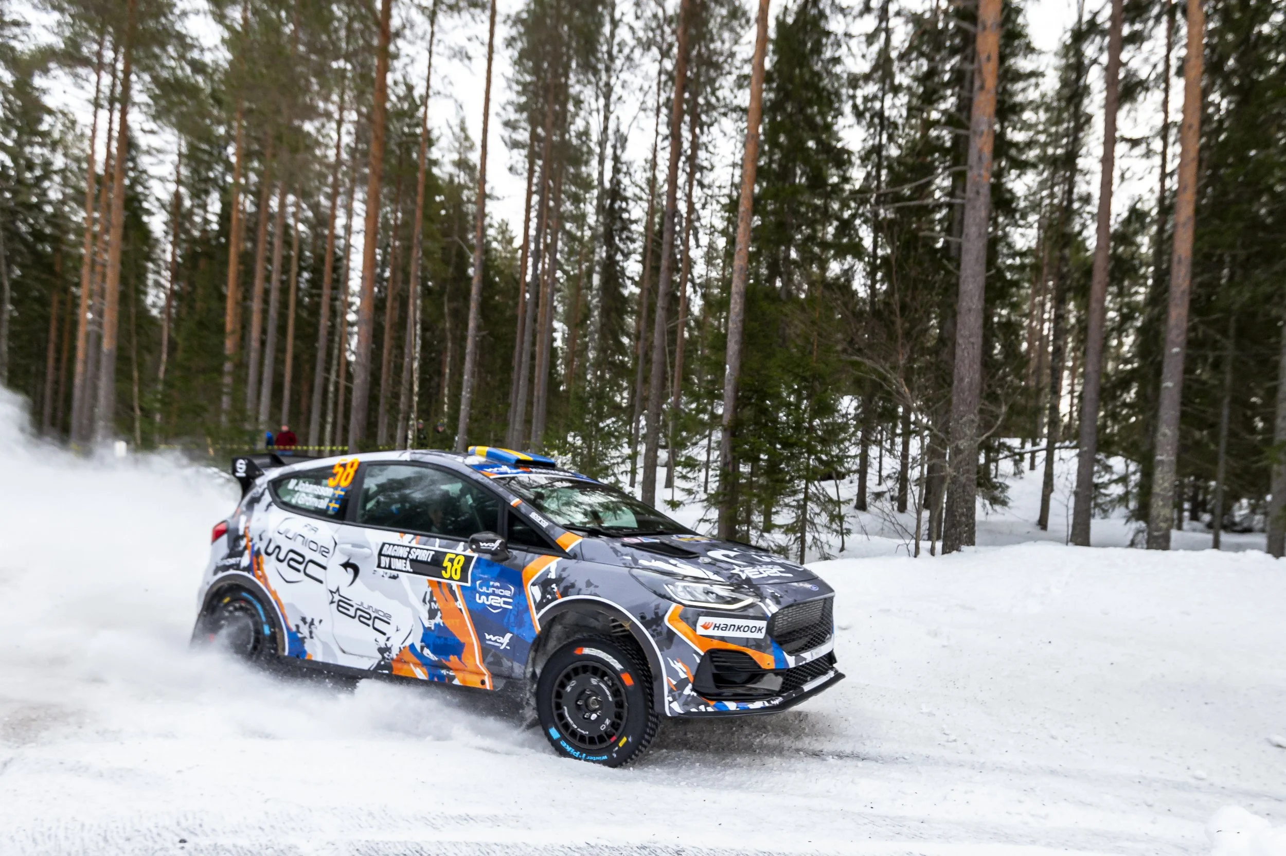 A rally car driving on a snow-covered course through a forest of tall pine trees, kicking up snow as it speeds along.