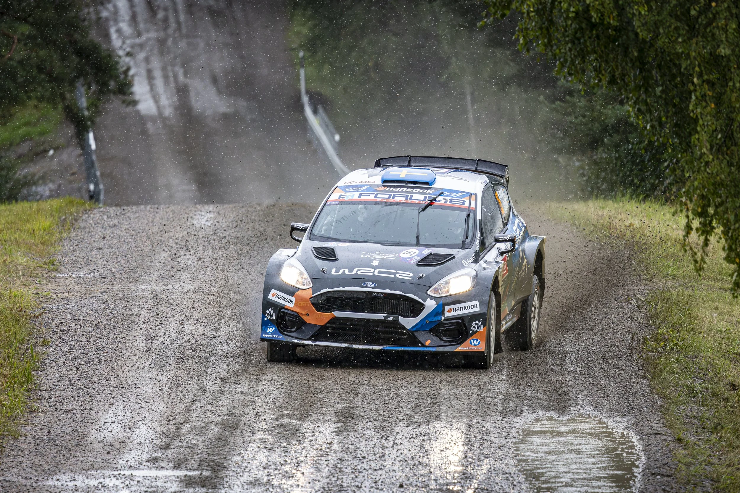 Rally car speeding on a gravel forest road, kicking up dust and water spray, surrounded by trees and greenery.