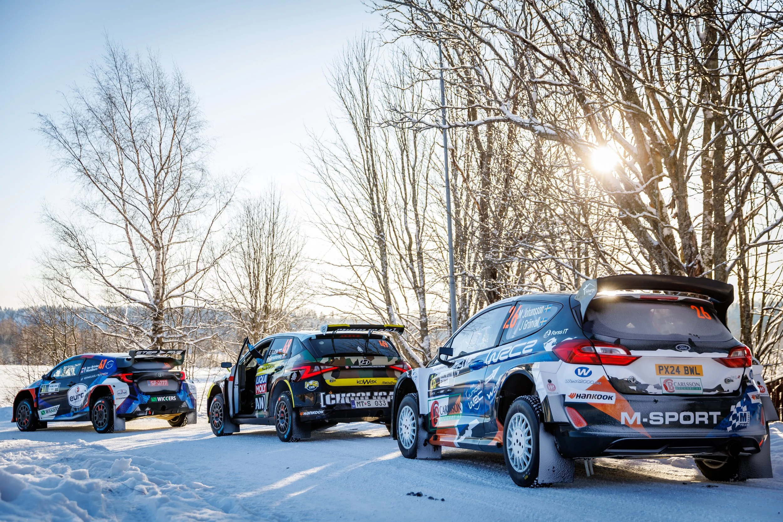 Line of rally cars parked on snow-covered ground with leafless trees and the sun in the background.