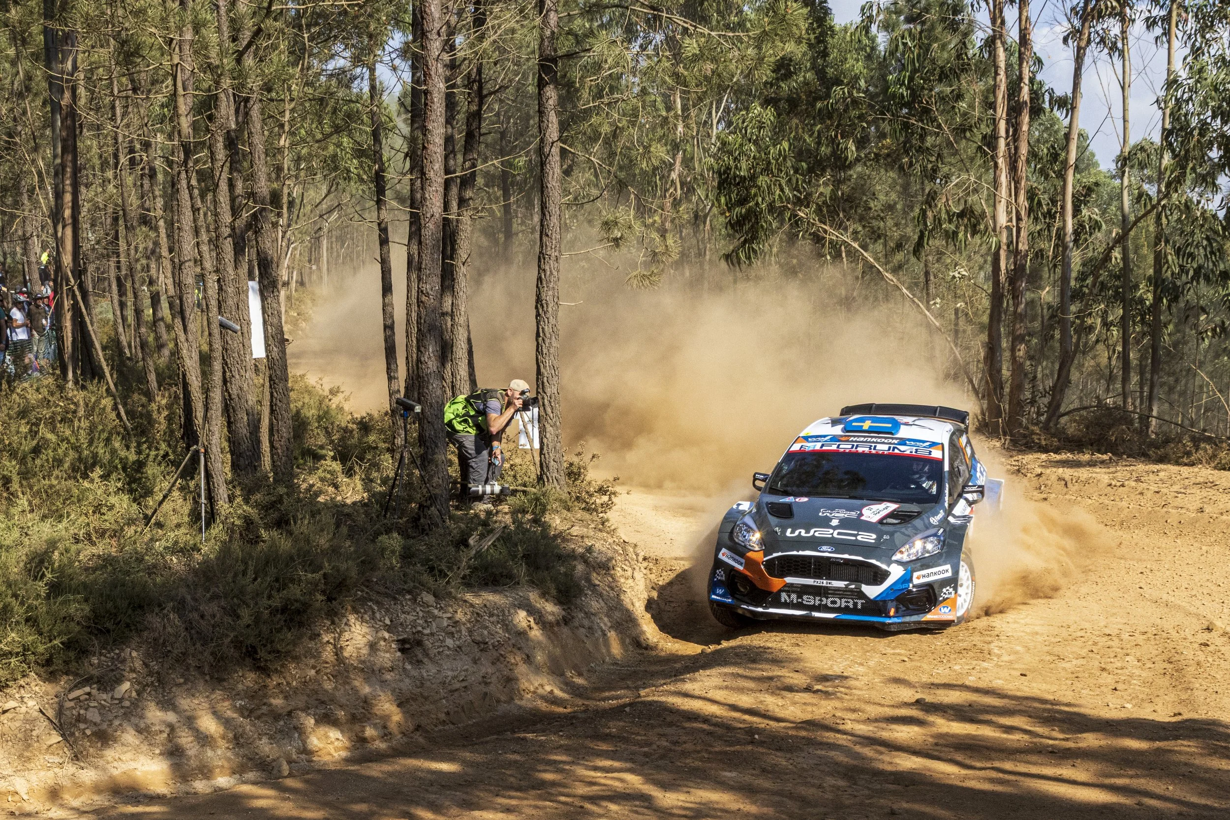 A rally car racing through a dirt forest trail, kicking up dust with photographers and spectators watching along the side.