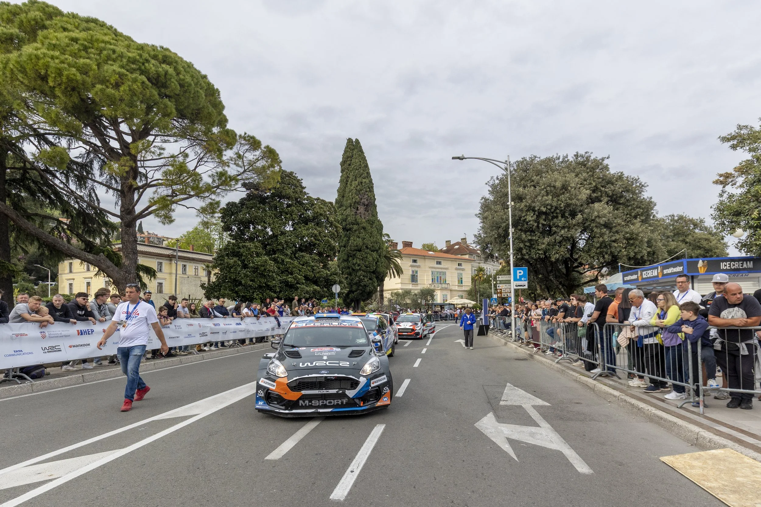 Race cars on a street with spectators lined up behind barriers, trees in the background, overcast sky, and people watching the event.