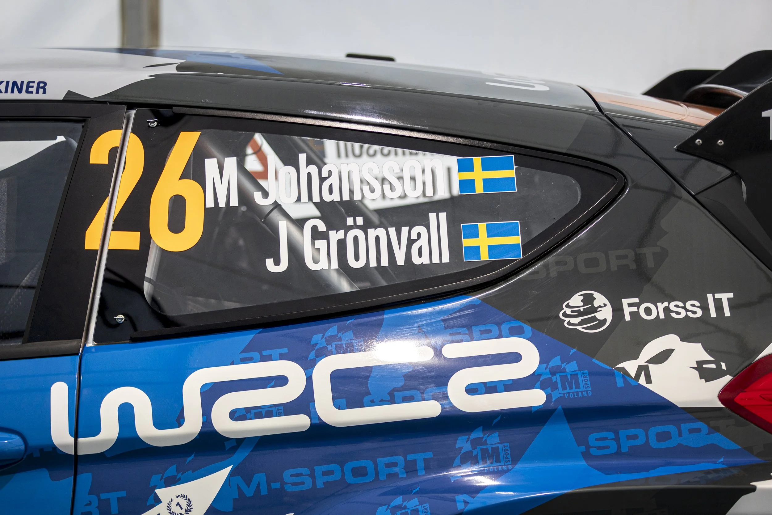 Close-up of a race car's window with the names M. Johansson and J. Grönvall, both Swedish flags, and the number 26.