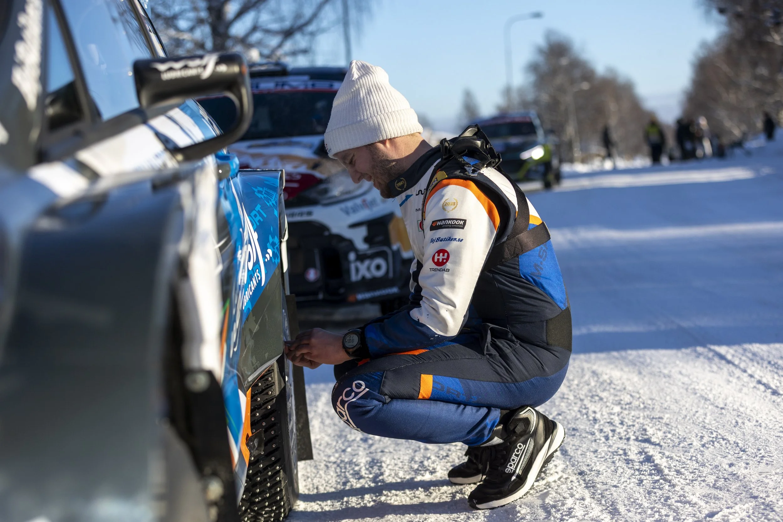 A race car driver in racing gear kneeling and working on a rally car on a snowy landscape, with other vehicles and people in the background during daylight.