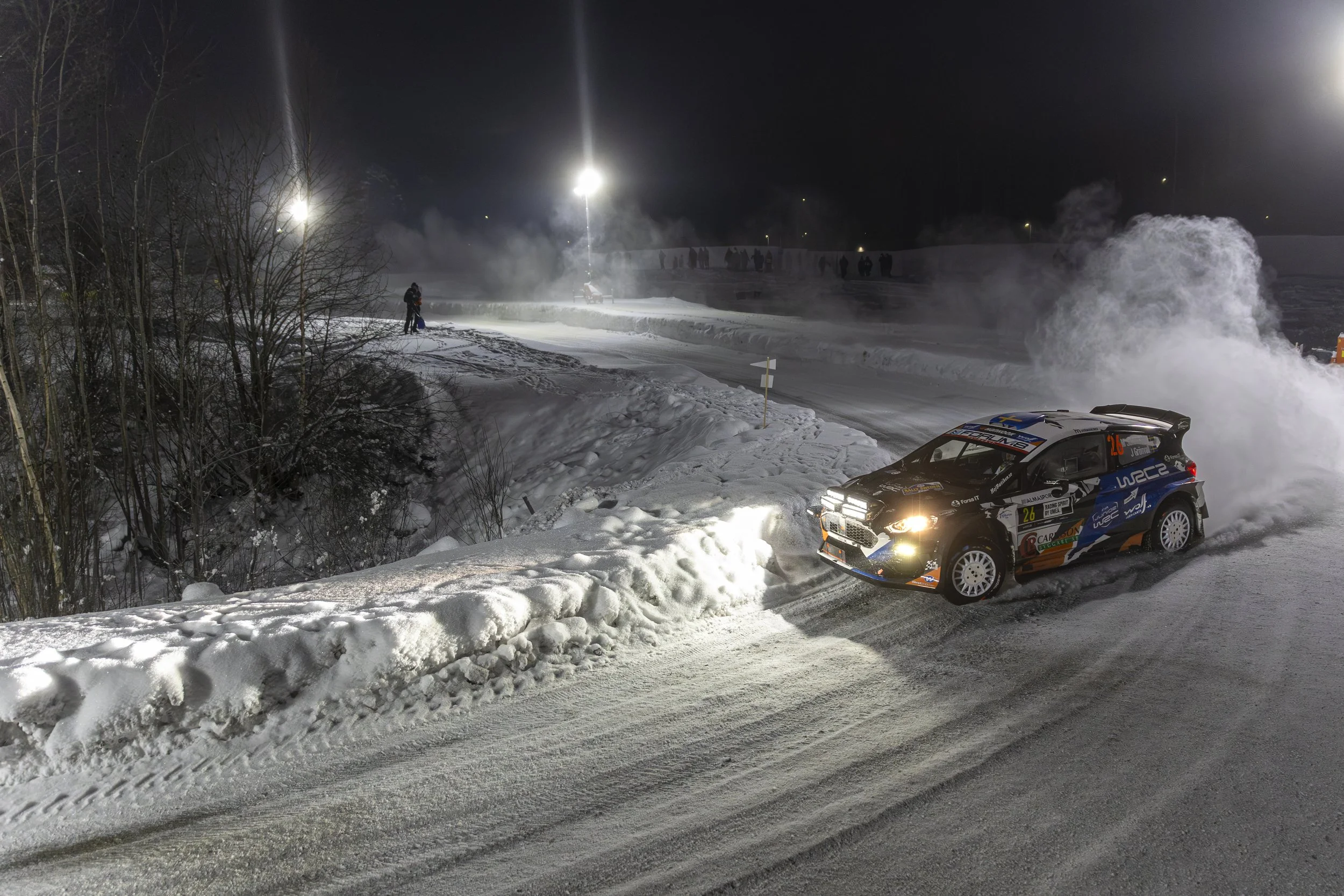 A rally car speeding on a snowy night, creating a spray of snow behind it, with spectators watching in the background and bright lights illuminating the scene.