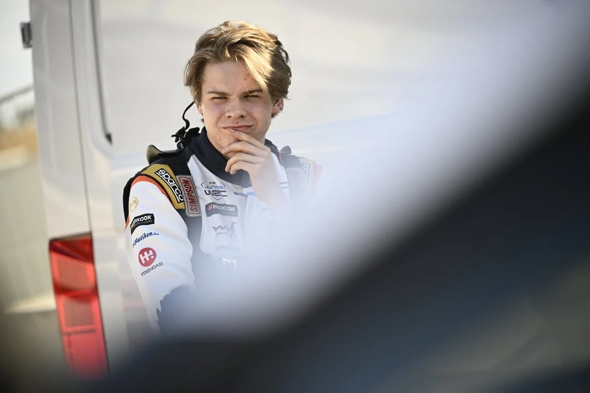 A young male race car driver with blonde hair, wearing a white racing suit with various sponsor logos, stands next to a vehicle, with a thoughtful expression and hand on his chin