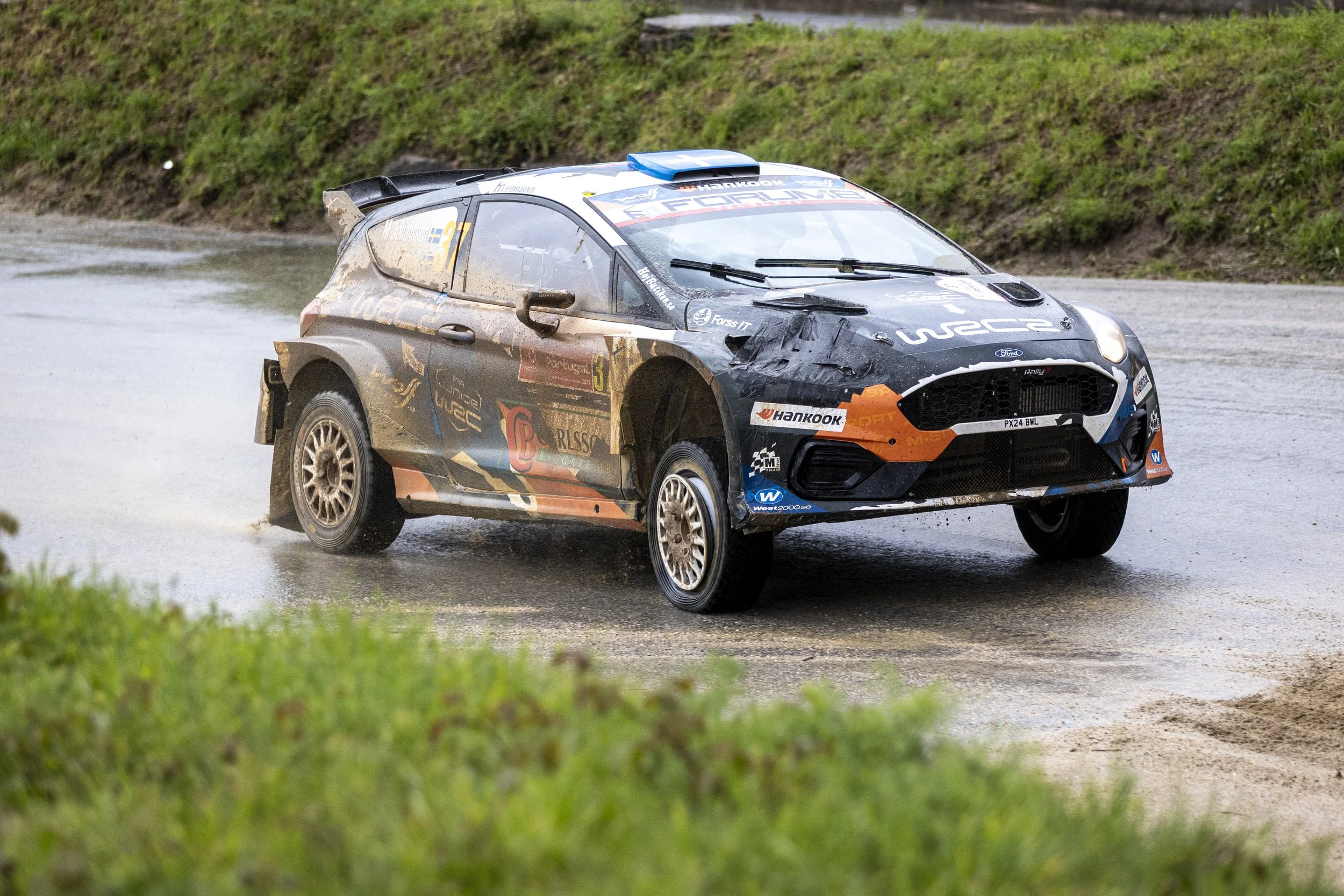 A rally car driving through a wet, muddy course with grass on each side.