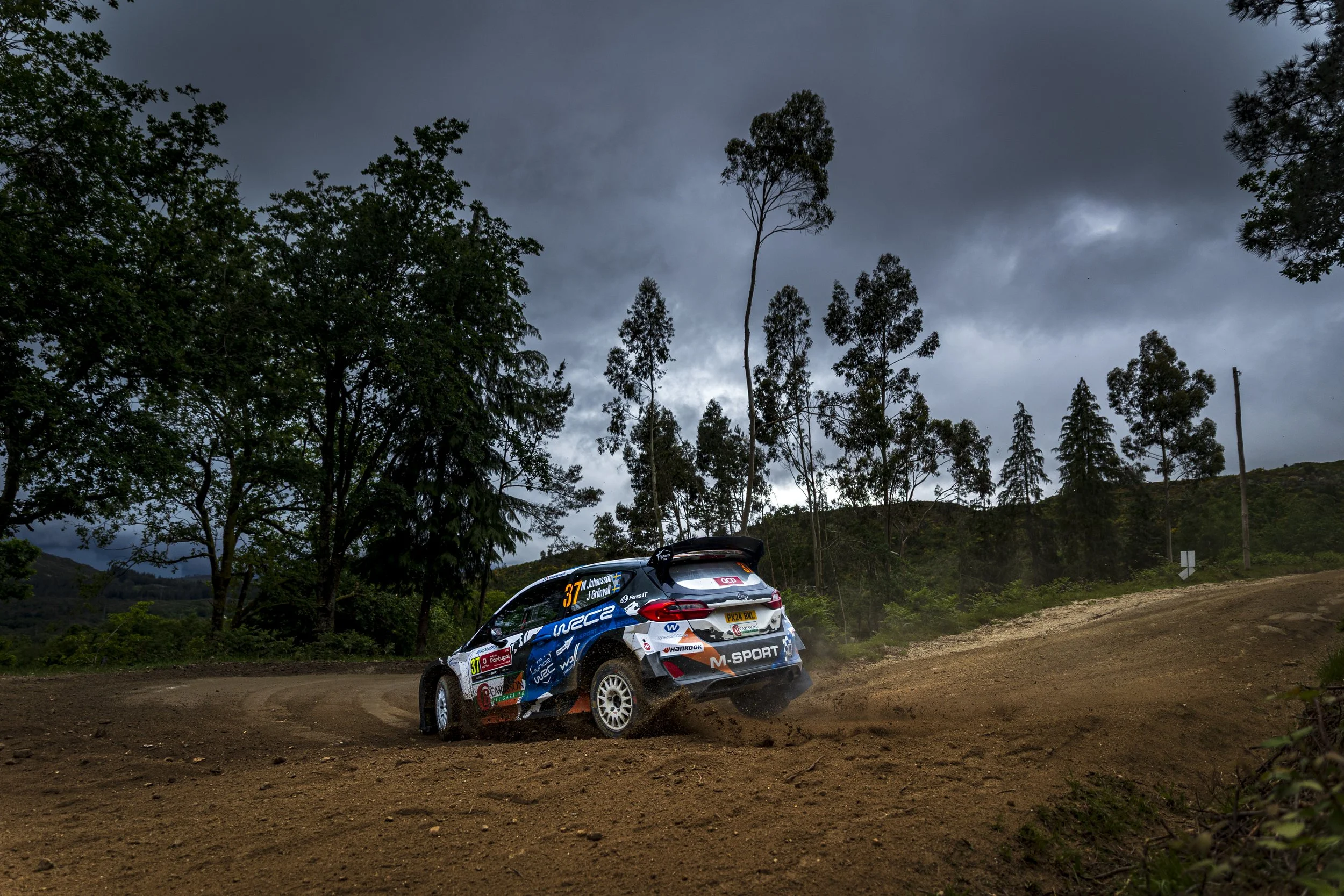 Rally car racing on an unpaved dirt road surrounded by trees and dark, cloudy sky.
